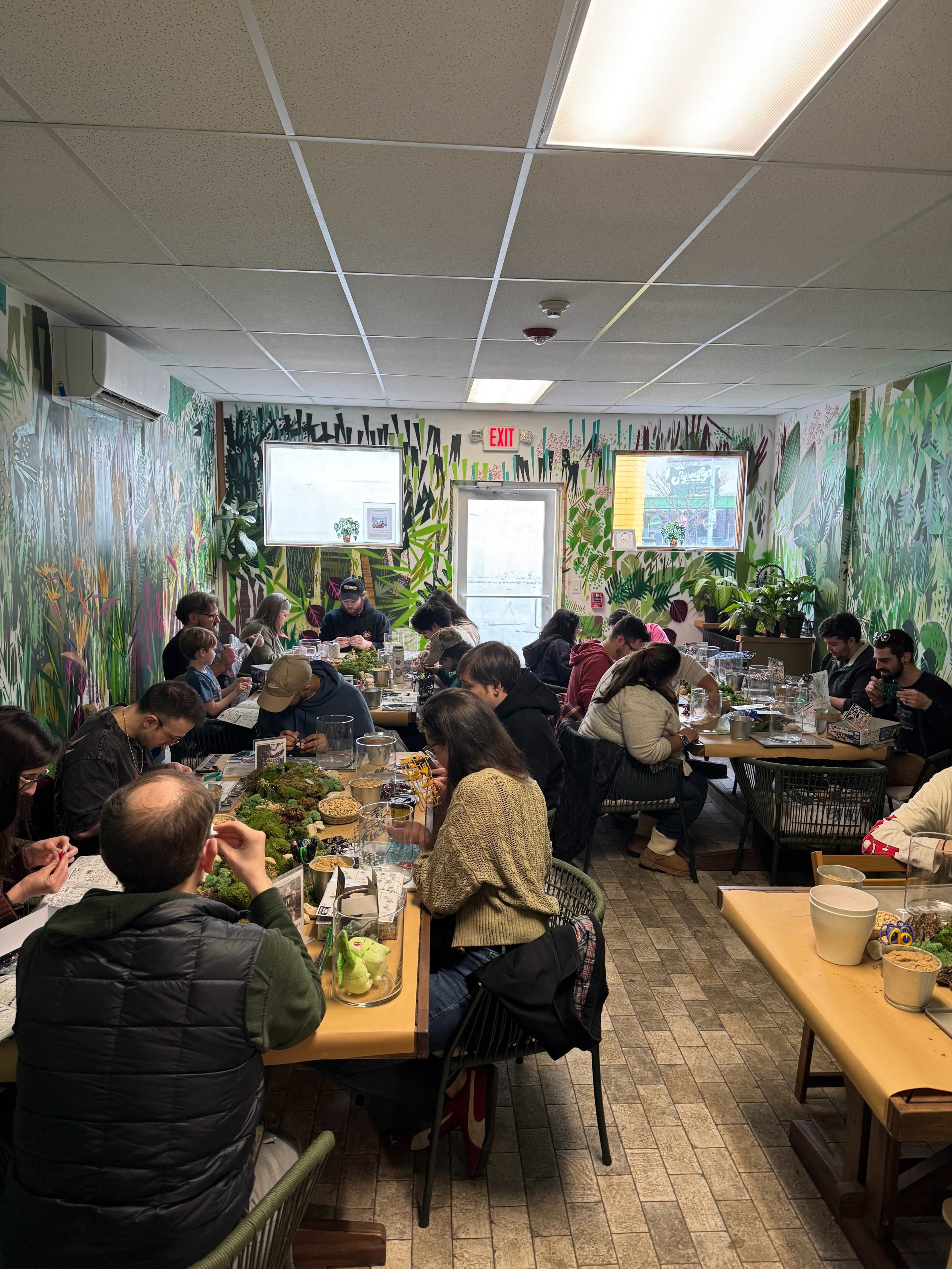 A busy café features a lively atmosphere where patrons are seated at multiple tables, enjoying their meals amidst vibrant green wall murals.
