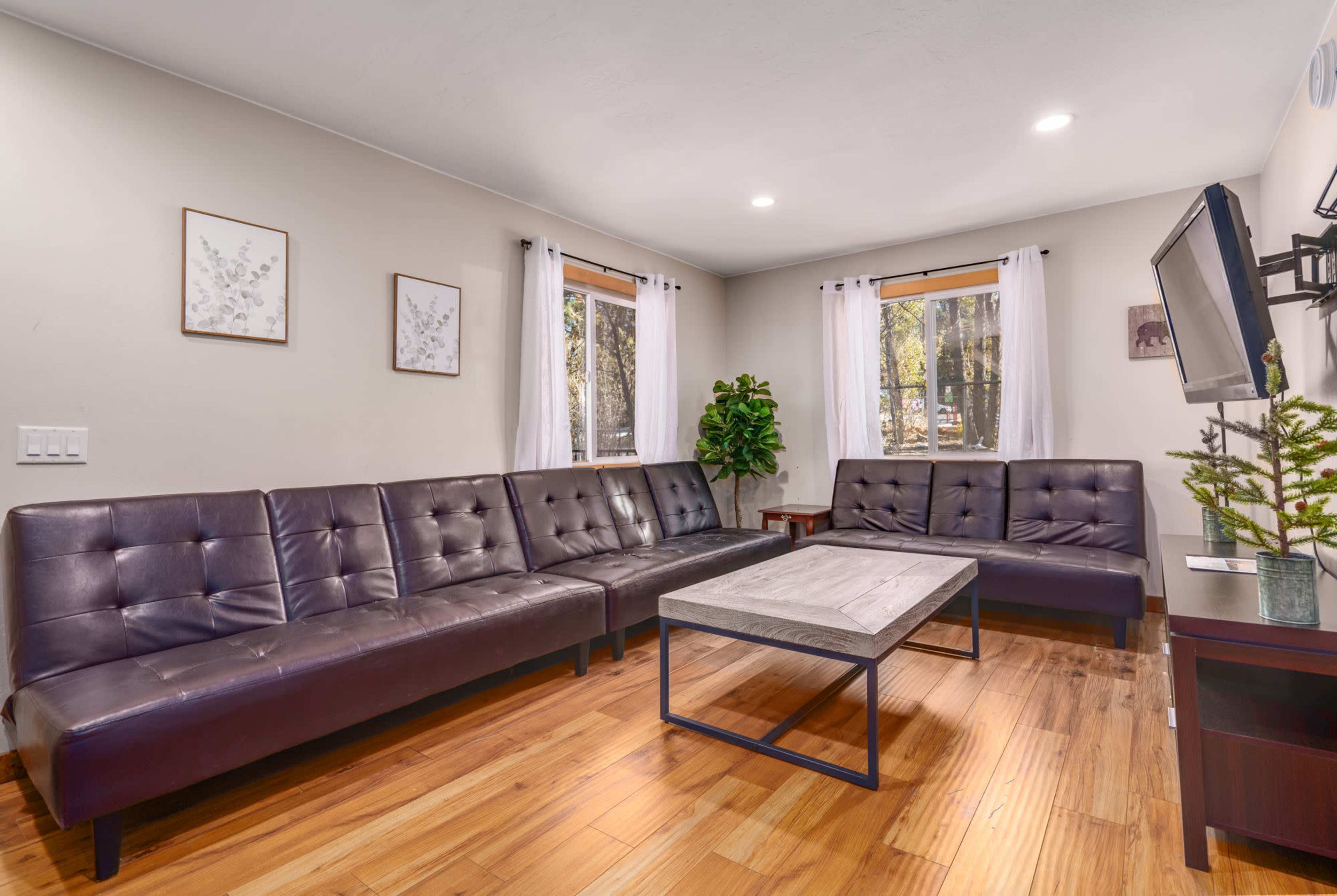 The living room features a large, dark leather sectional sofa arranged around a wooden coffee table, with large windows letting in natural light.
