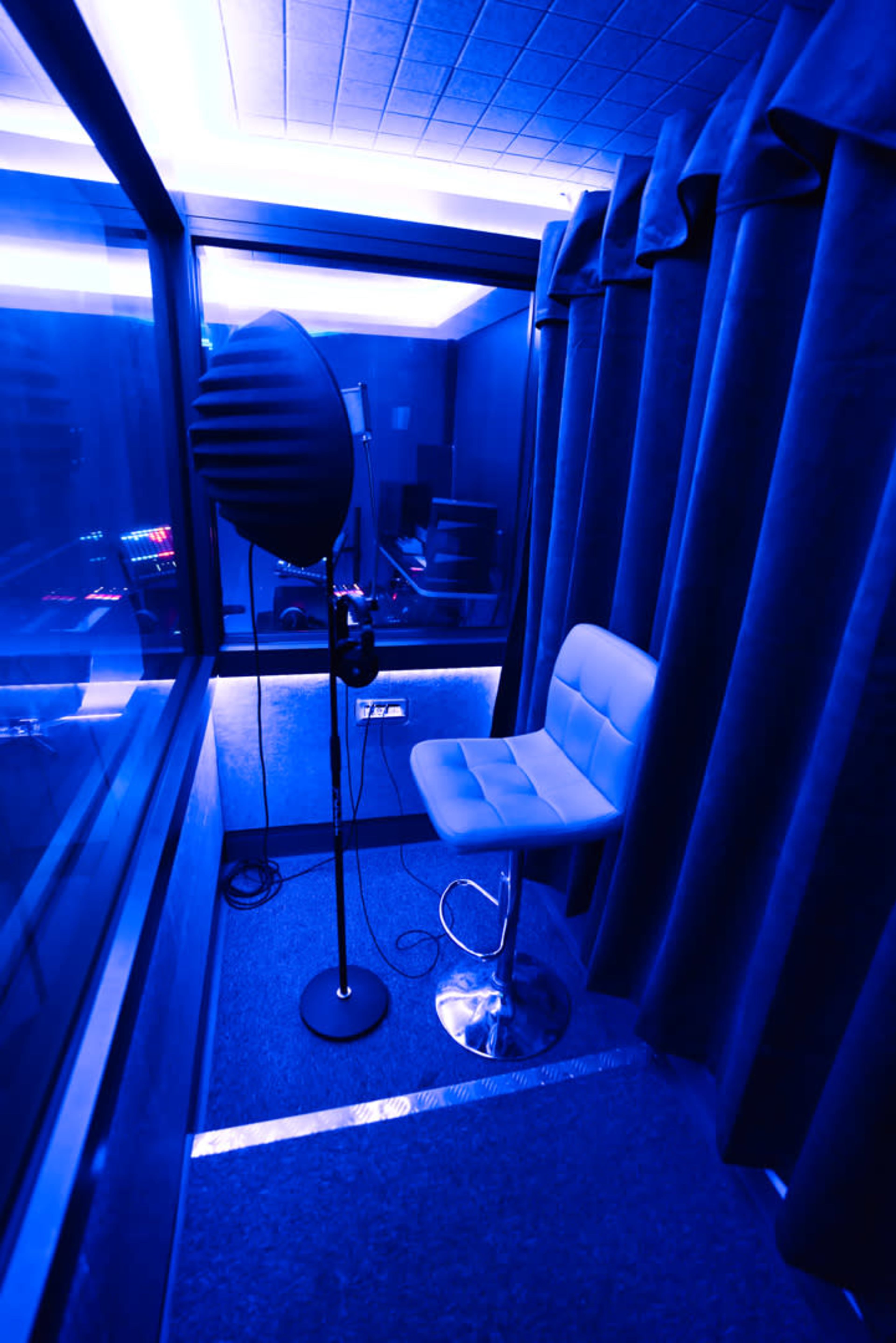 A soundproof recording booth features a microphone on a stand, a padded stool, and blue lighting.