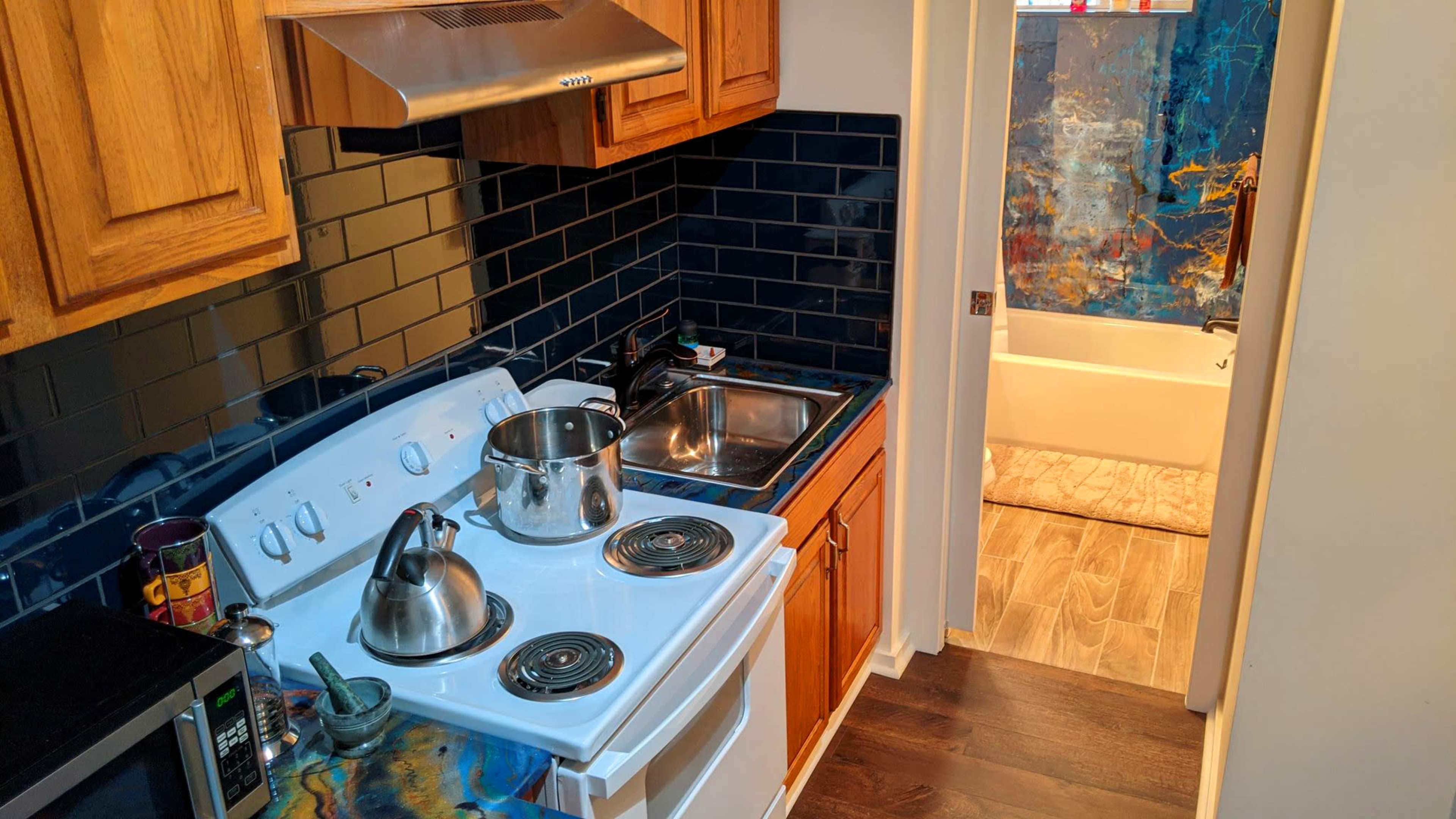 A compact kitchen features a stove, sink, and a kettle, with a bathroom visible in the background.