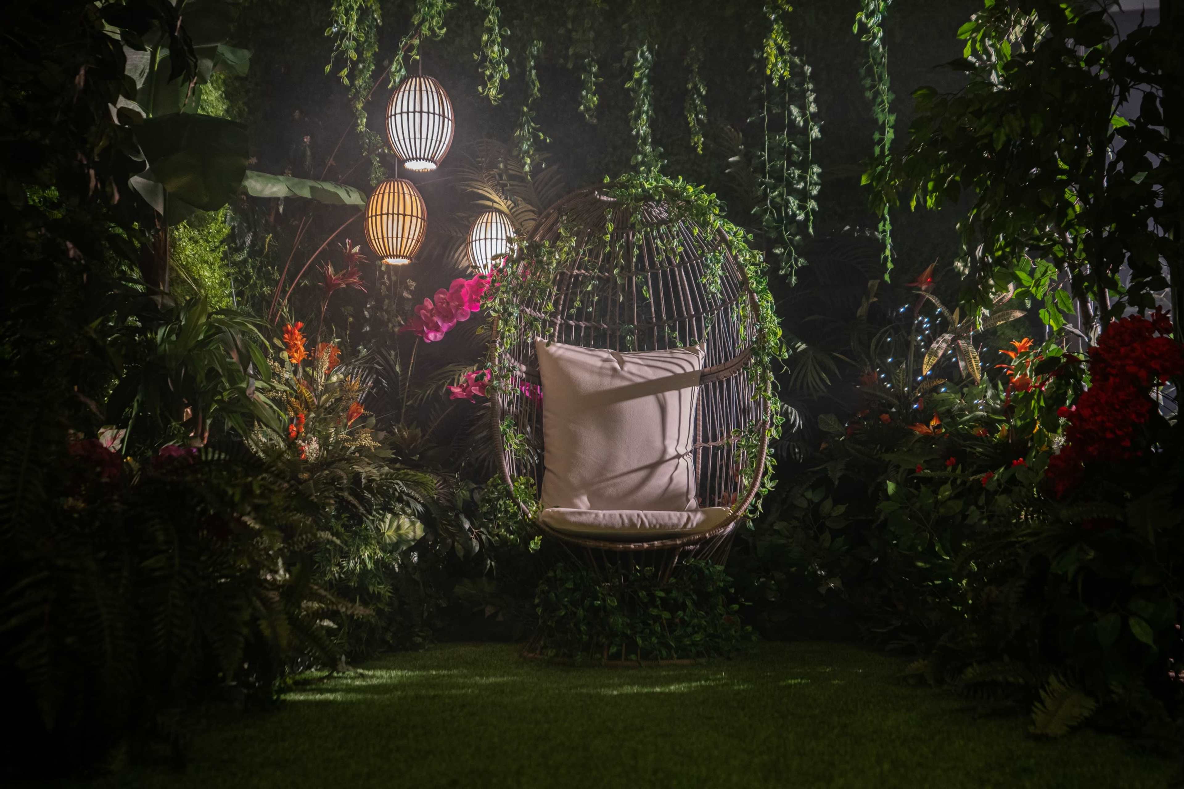 A hanging chair is surrounded by lush greenery and colorful plants, illuminated by decorative lamps in a serene garden setting.