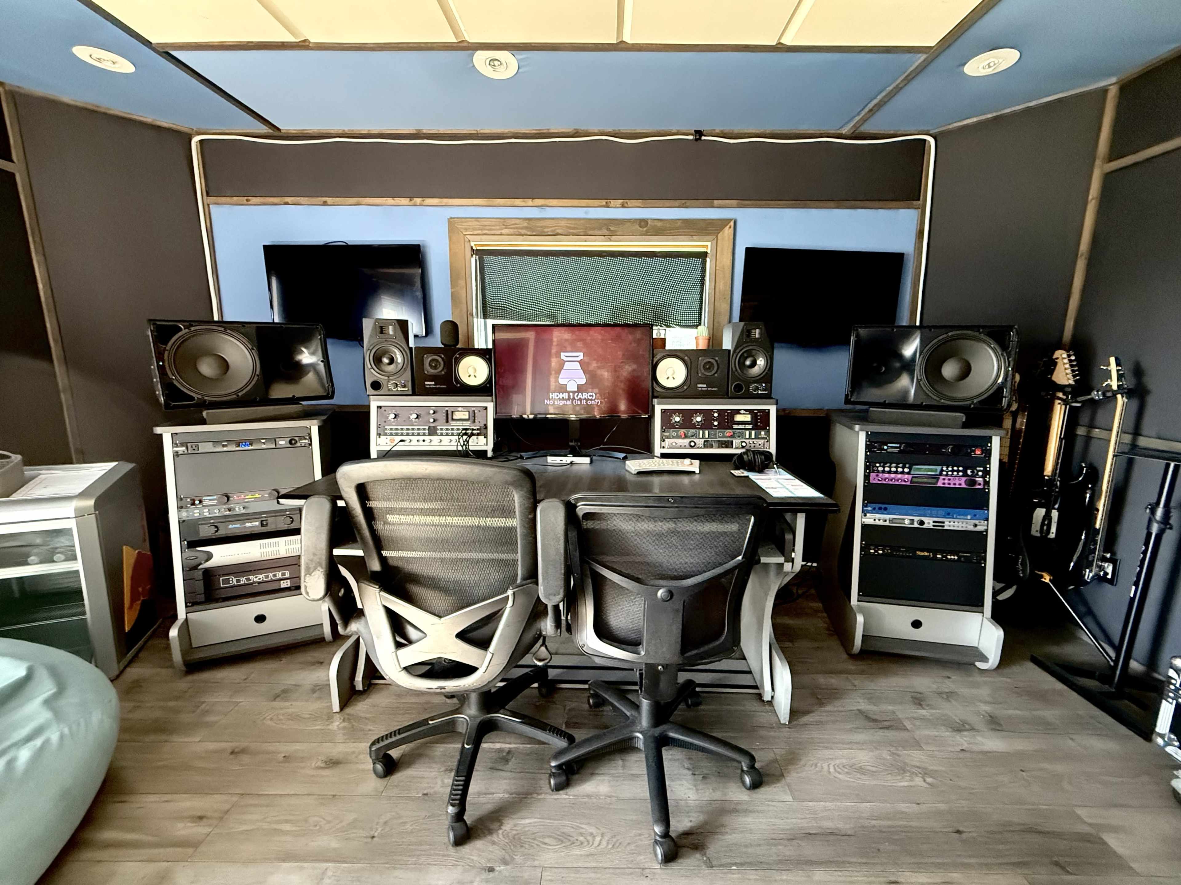 The image shows a music production studio featuring two office chairs positioned in front of a digital audio workstation, surrounded by speakers and various audio equipment.