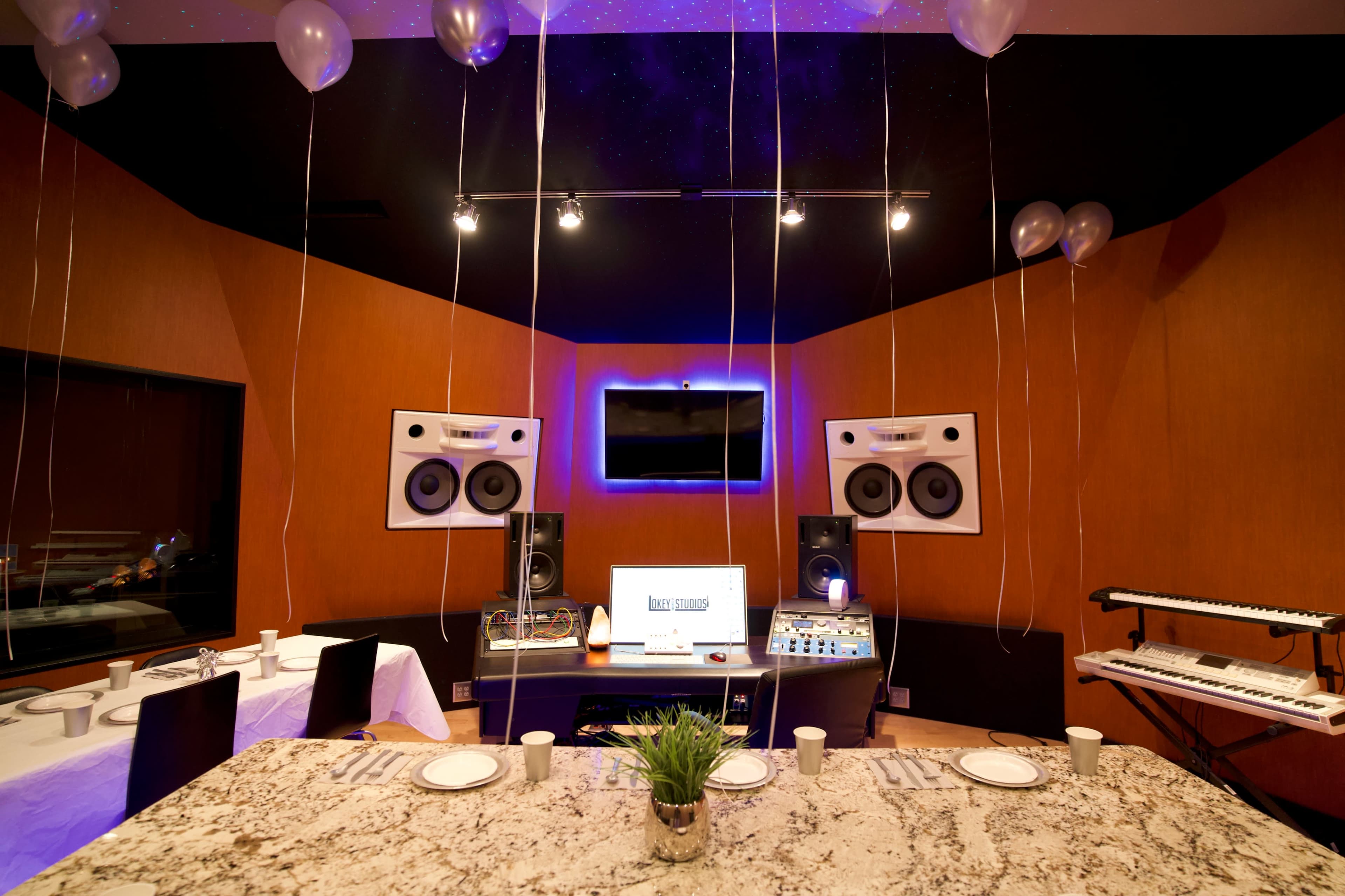 A music studio is set up with a table and white decorations, featuring two large speakers, a mixing console, and balloons hanging from the ceiling.