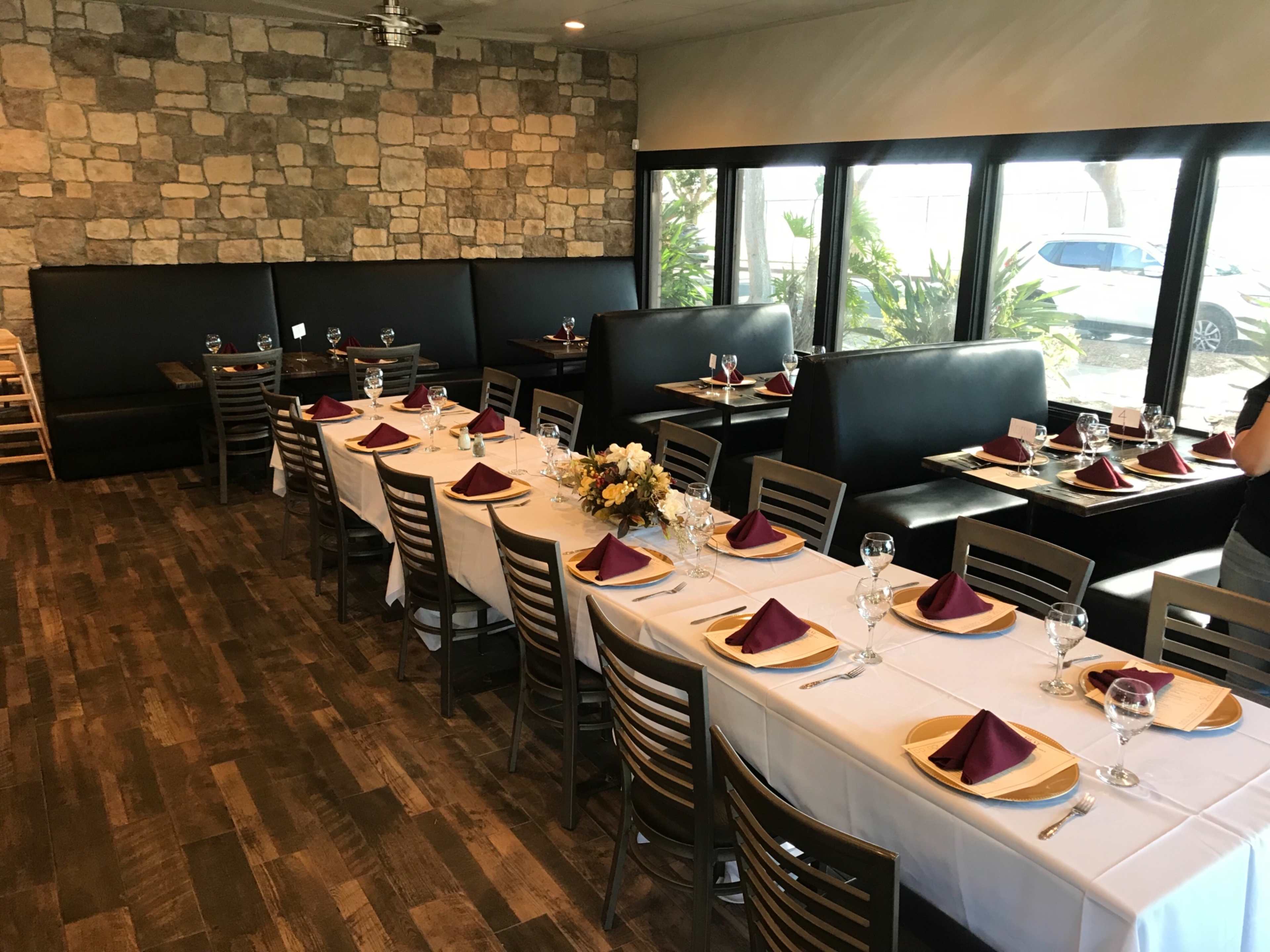 A dining area with a long table set for a meal, surrounded by smaller tables, and features modern black booths and large windows.