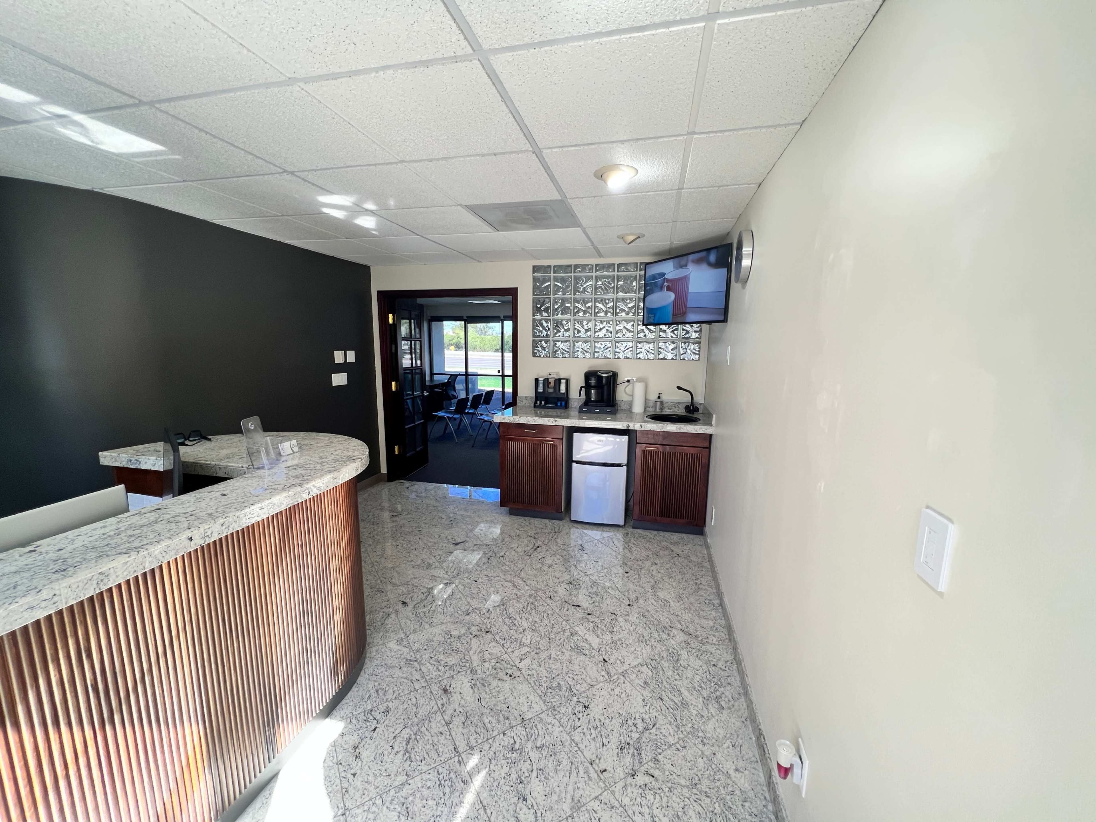 A modern reception area with a curved front desk, a coffee station, a refrigerator, and a large window overlooking a meeting space.