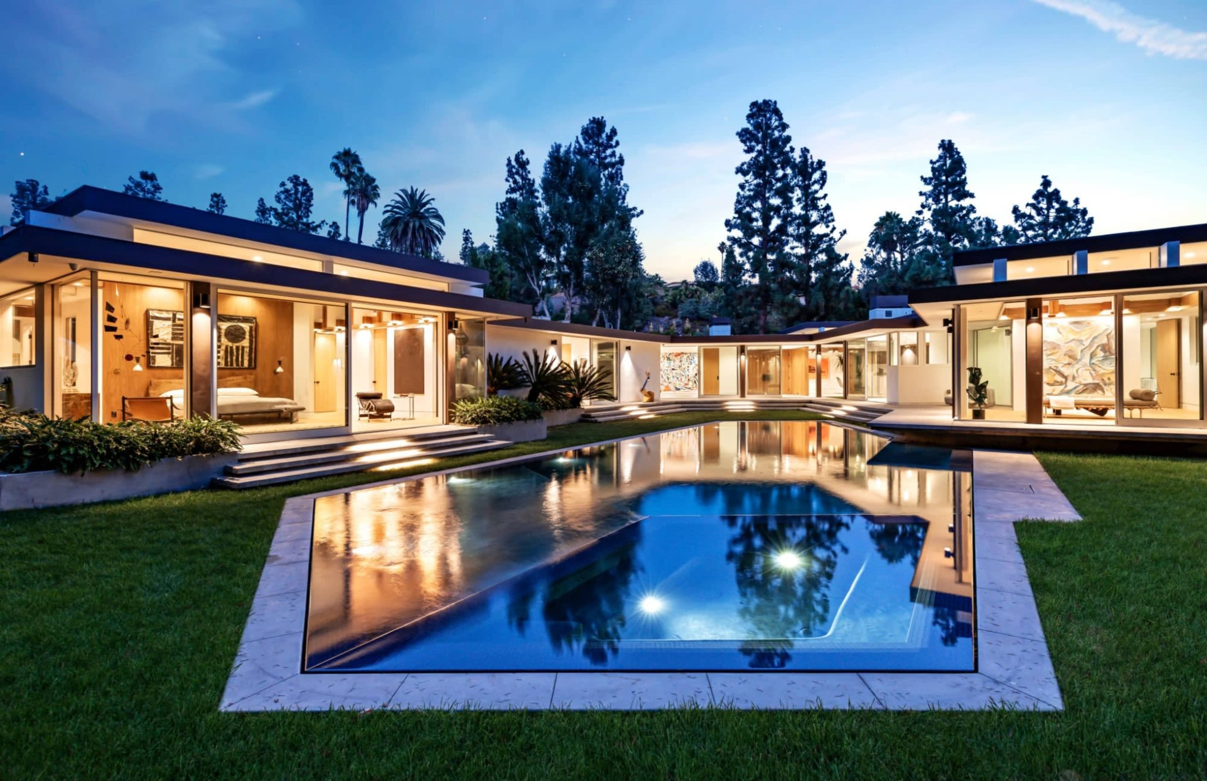 A modern house with an expansive pool is surrounded by landscaped gardens and tall trees under a twilight sky.