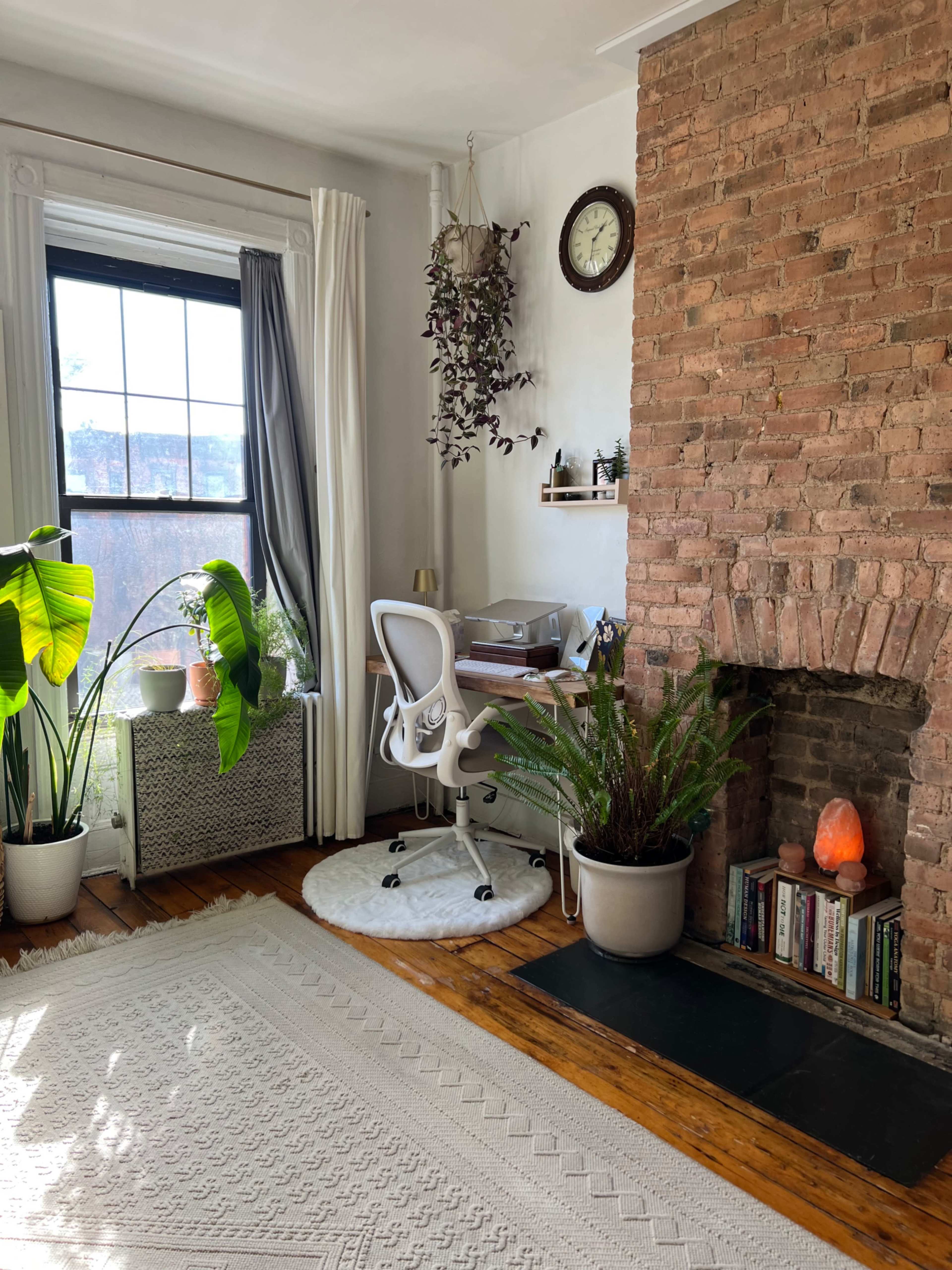 The image shows a cozy work area in a room with a large window, featuring a desk next to a brick fireplace, a plant in the foreground, and a soft rug on the floor.