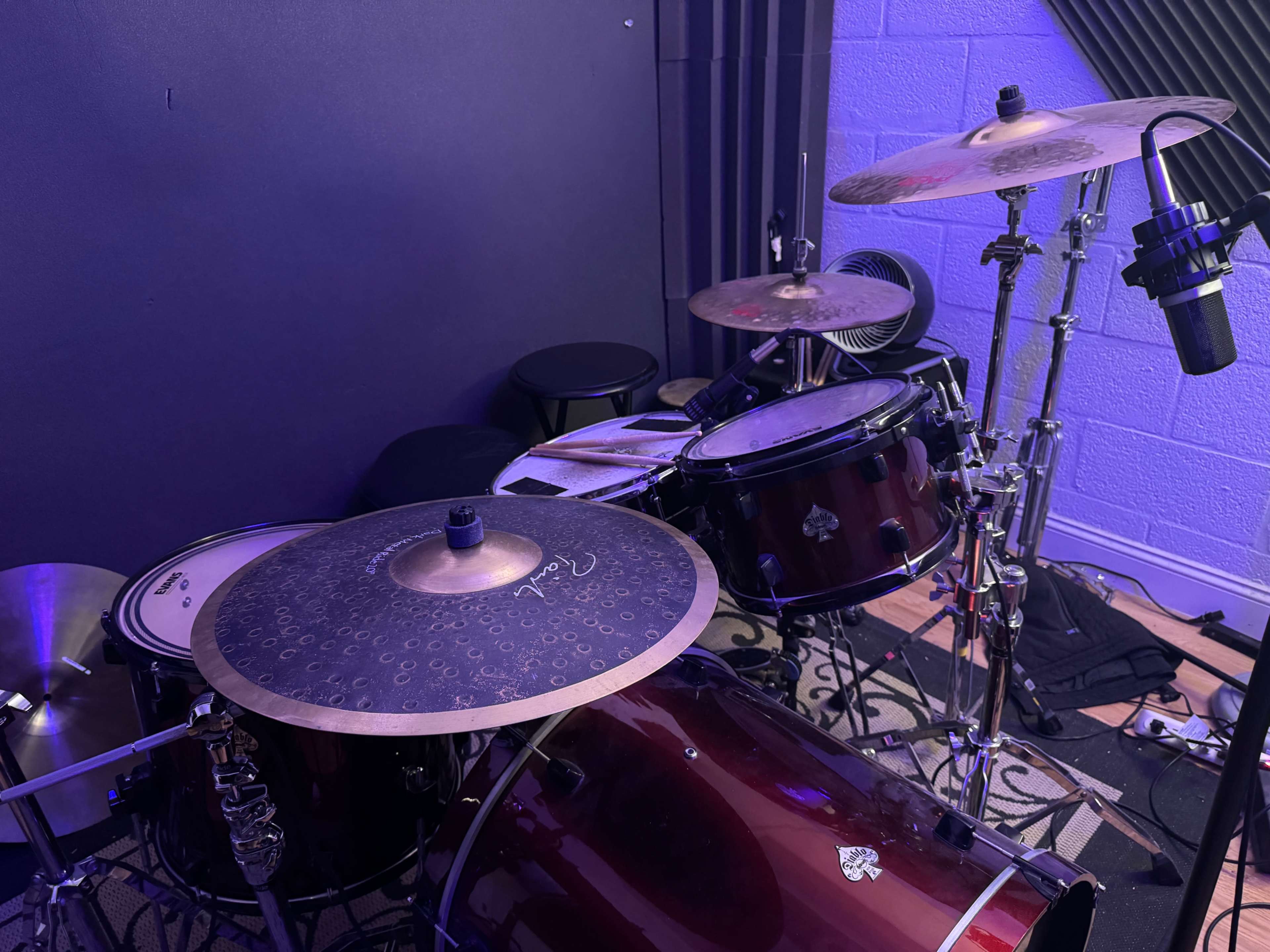 A drum set with multiple drums and cymbals is positioned in a music studio, accompanied by a microphone and a stool.