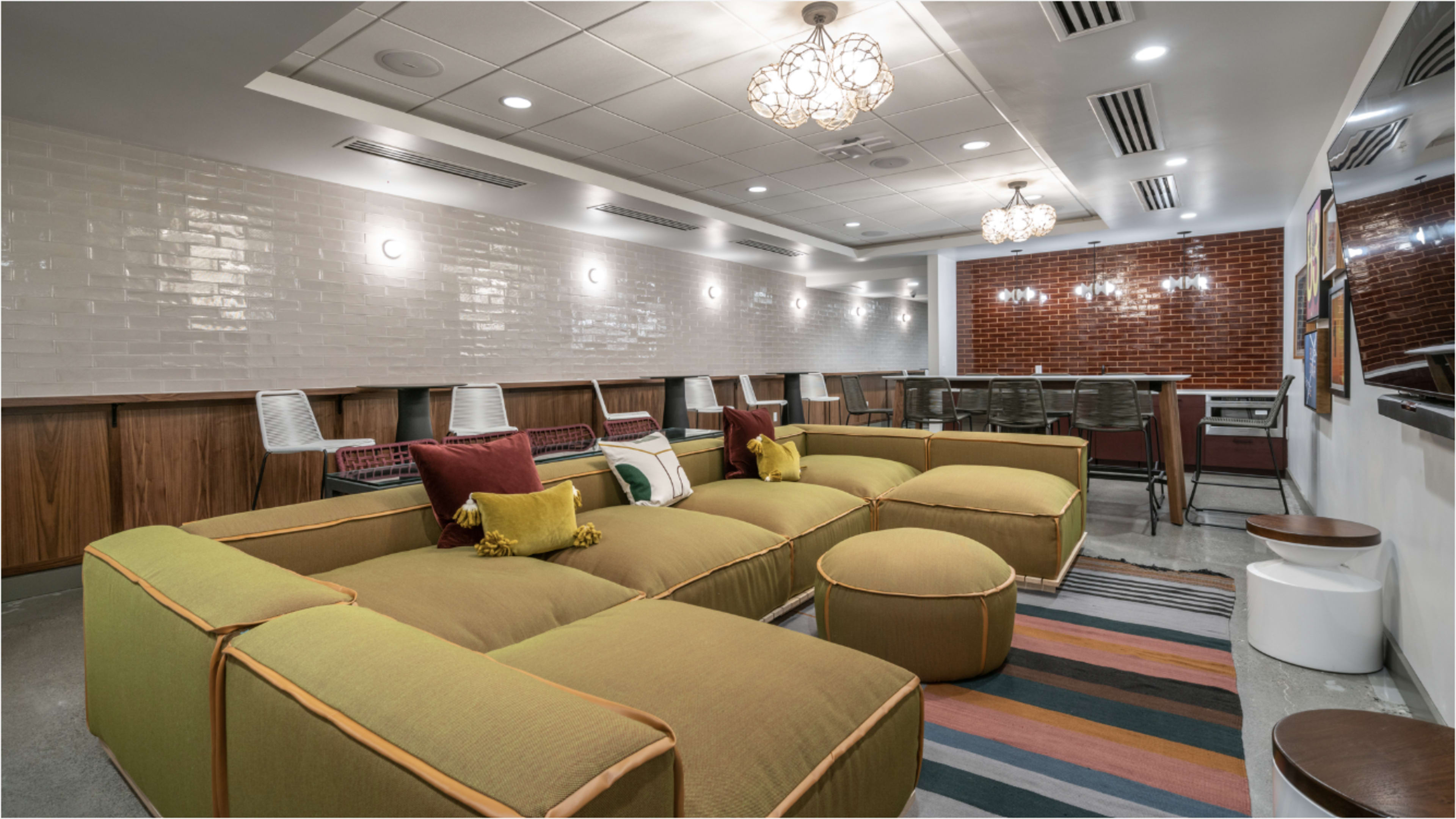 The image shows a modern lounge area featuring a large, green sectional sofa, circular poufs, and a long dining table in a well-lit room with decorative wall lighting.