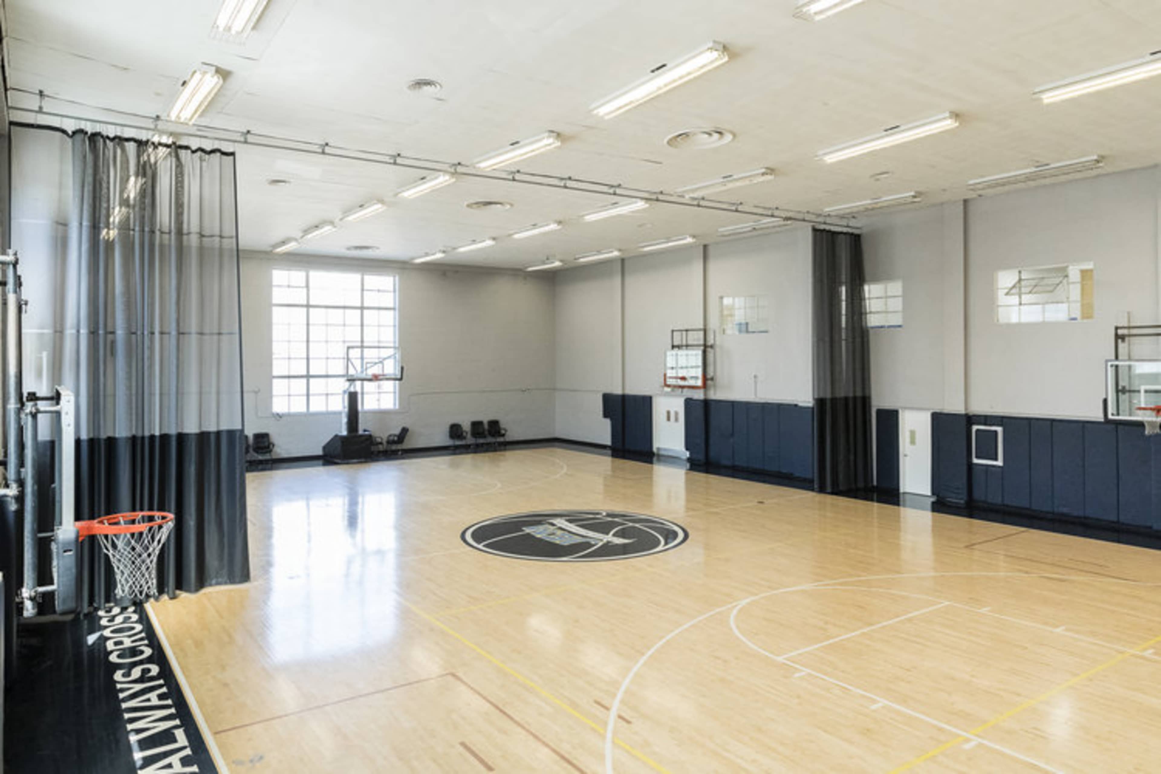 A spacious indoor basketball court with a polished wooden floor, basketball hoops on both ends, and a curtain dividing the court area.