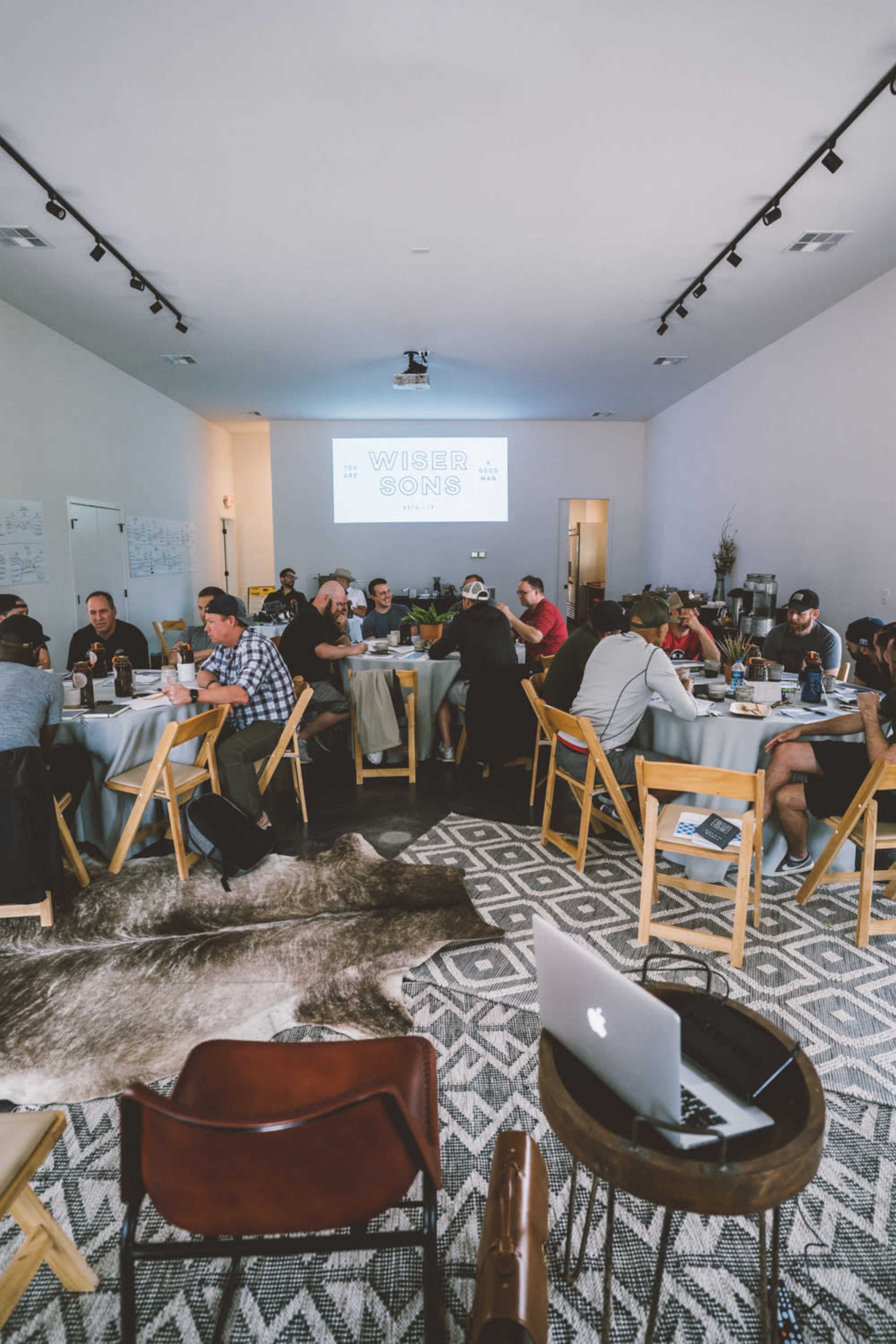 A group of people sits around tables in a modern meeting space with a large screen displaying the text "WIDER SONS."