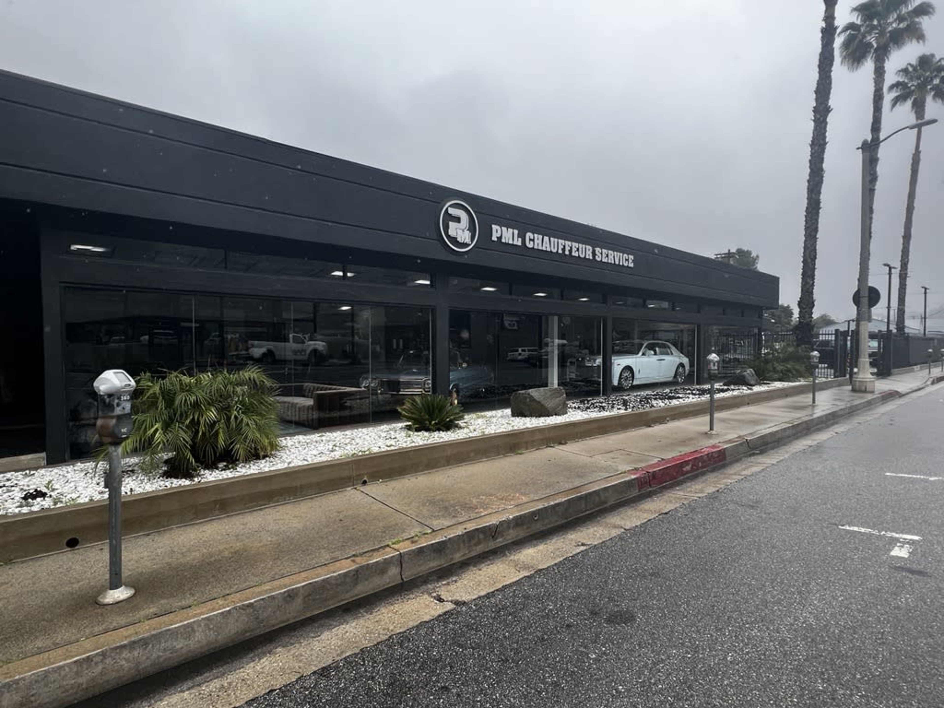 A modern building housing a chauffeur service, featuring large windows and palm trees in front, on a rainy day.