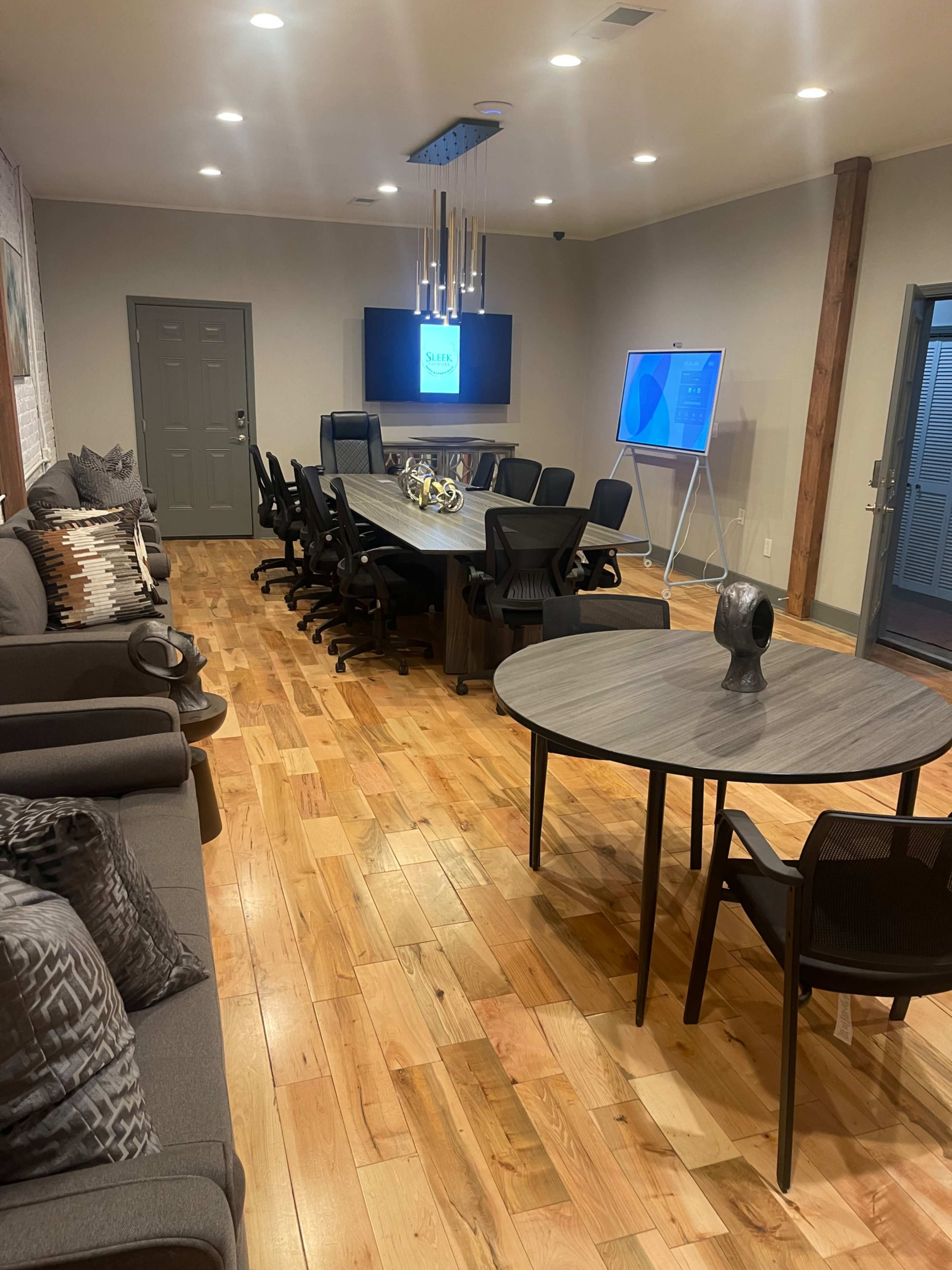 A modern conference room with a long table surrounded by chairs, a large screen on the wall, and a small round table with a decorative piece.