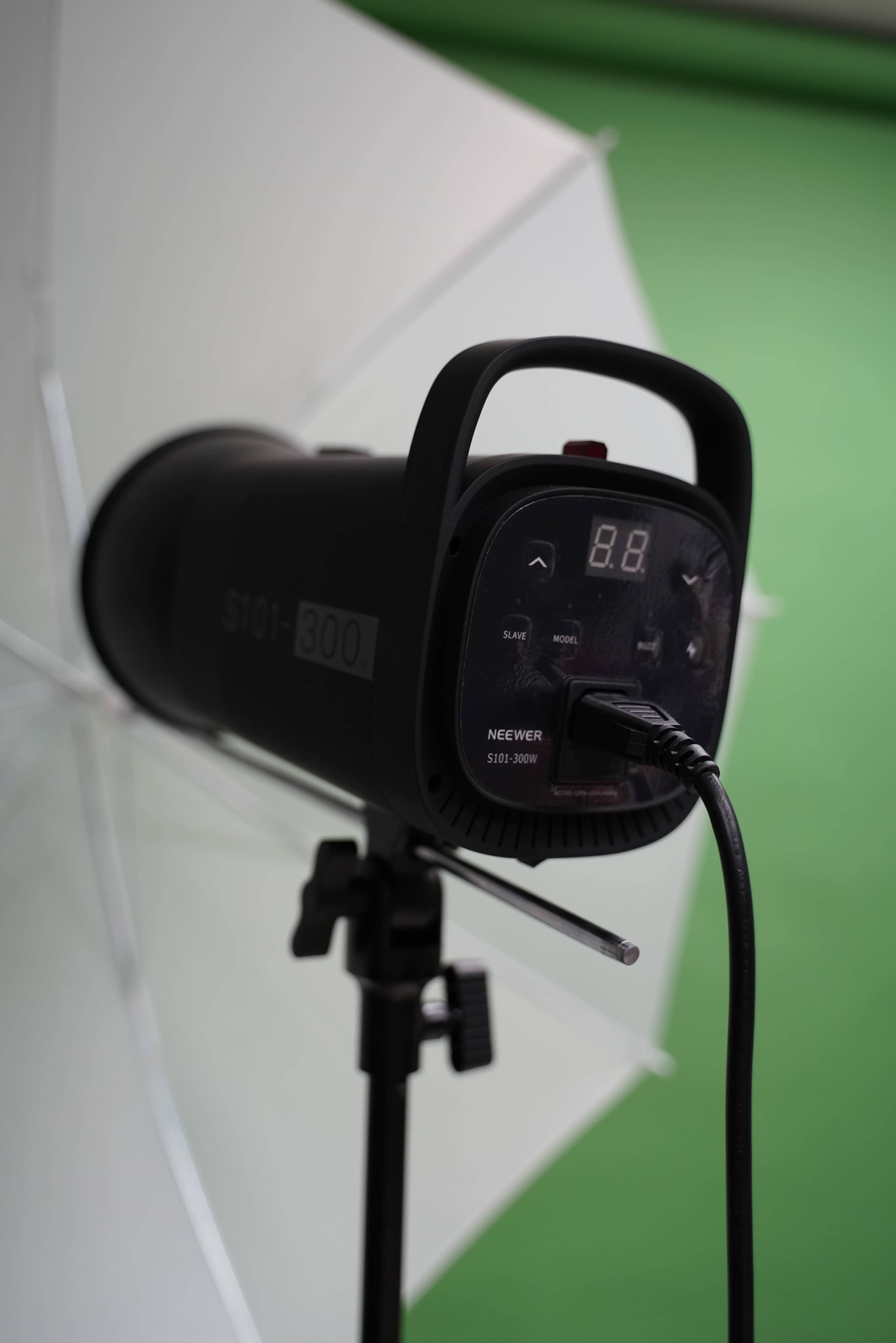 A Neewer S101-300 studio light is mounted on a stand, equipped with an umbrella and set against a green backdrop.