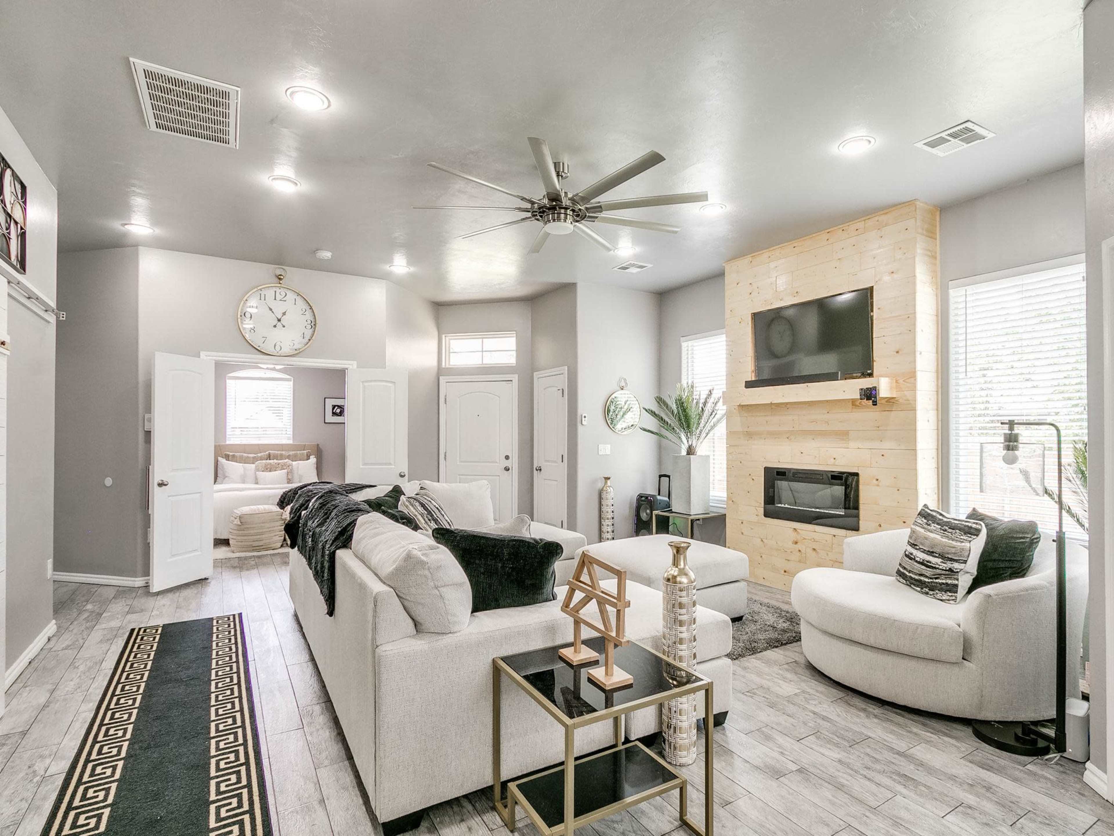 A modern living room with a light color palette, featuring a sectional sofa, two armchairs, a coffee table, a mounted television, and a decorative fireplace.