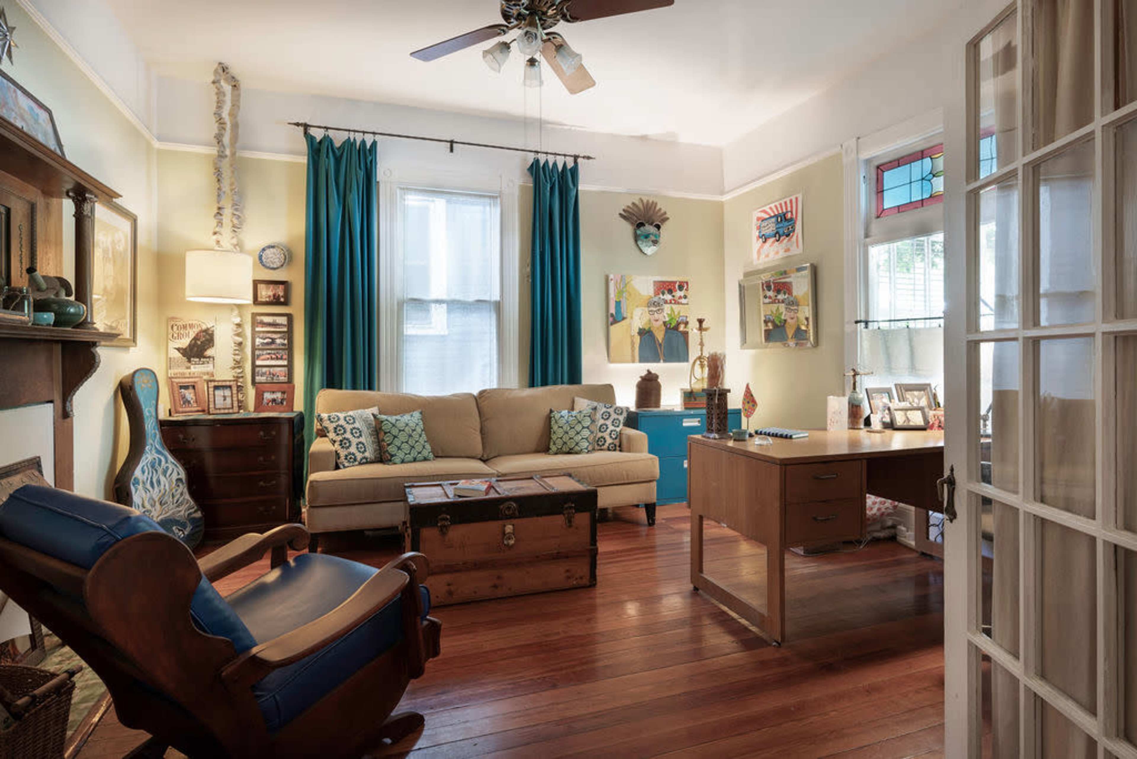 The image shows a cozy living room with a beige sofa, a wooden desk, and a blue cabinet, featuring decorative art on the walls and blue curtains.