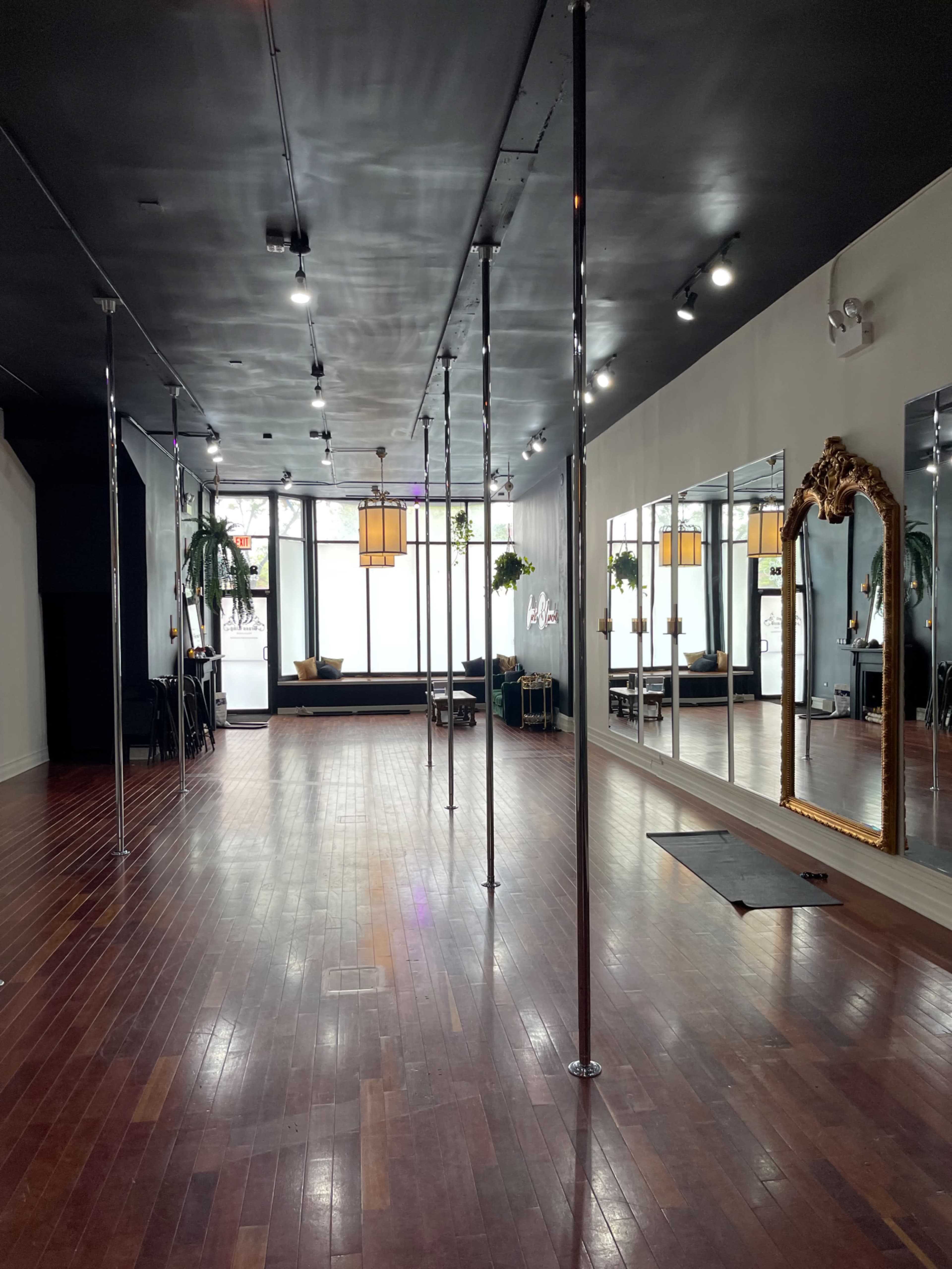 The image shows a spacious studio with polished wooden floors, several pole dance poles, large mirrors, and a large decorative mirror against one wall.