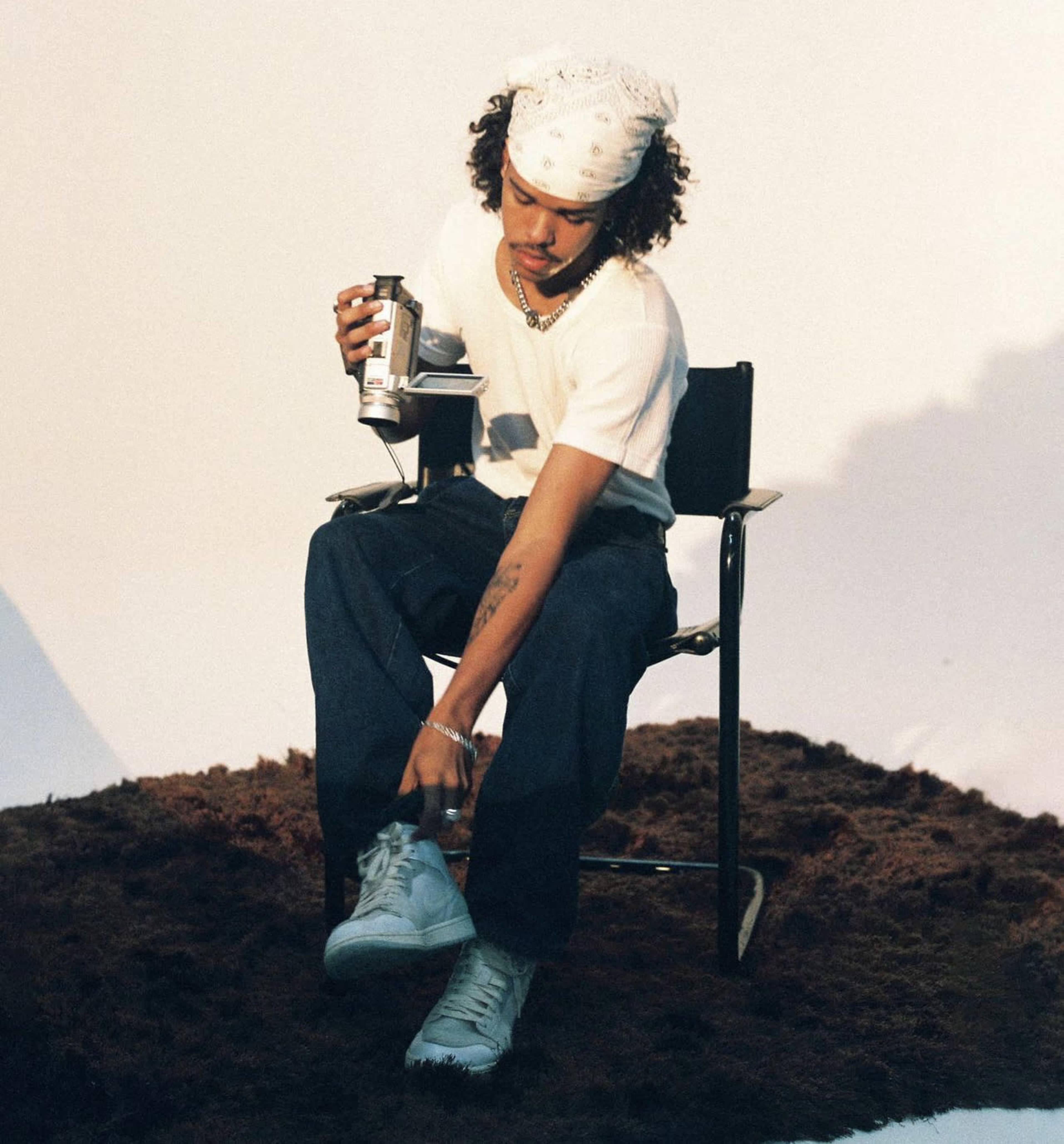 A person sitting on a chair adjusts their sneaker while holding a can and wearing a bandana.