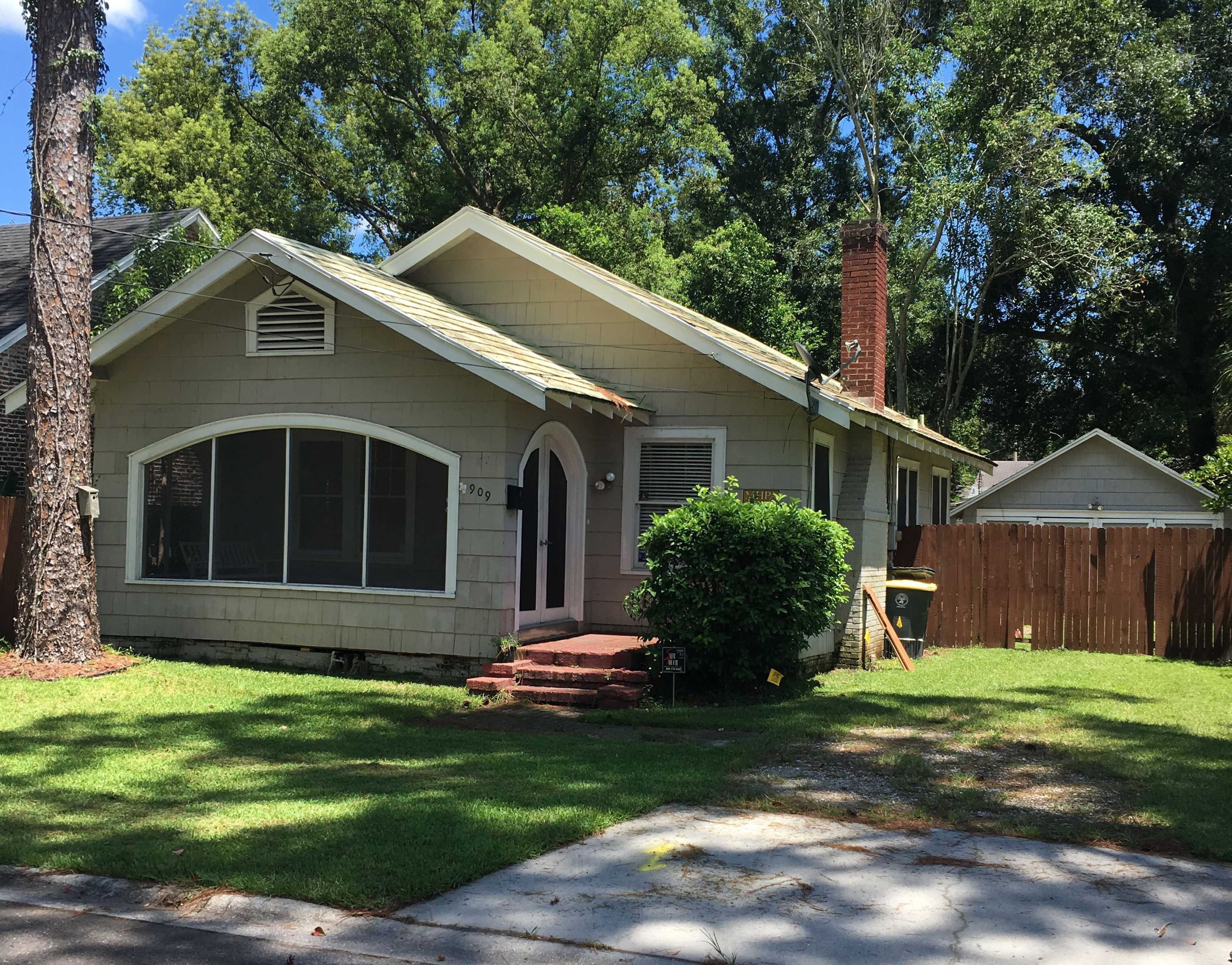 A single-story, light-colored house with a sloped roof and a front porch is surrounded by green grass and trees.