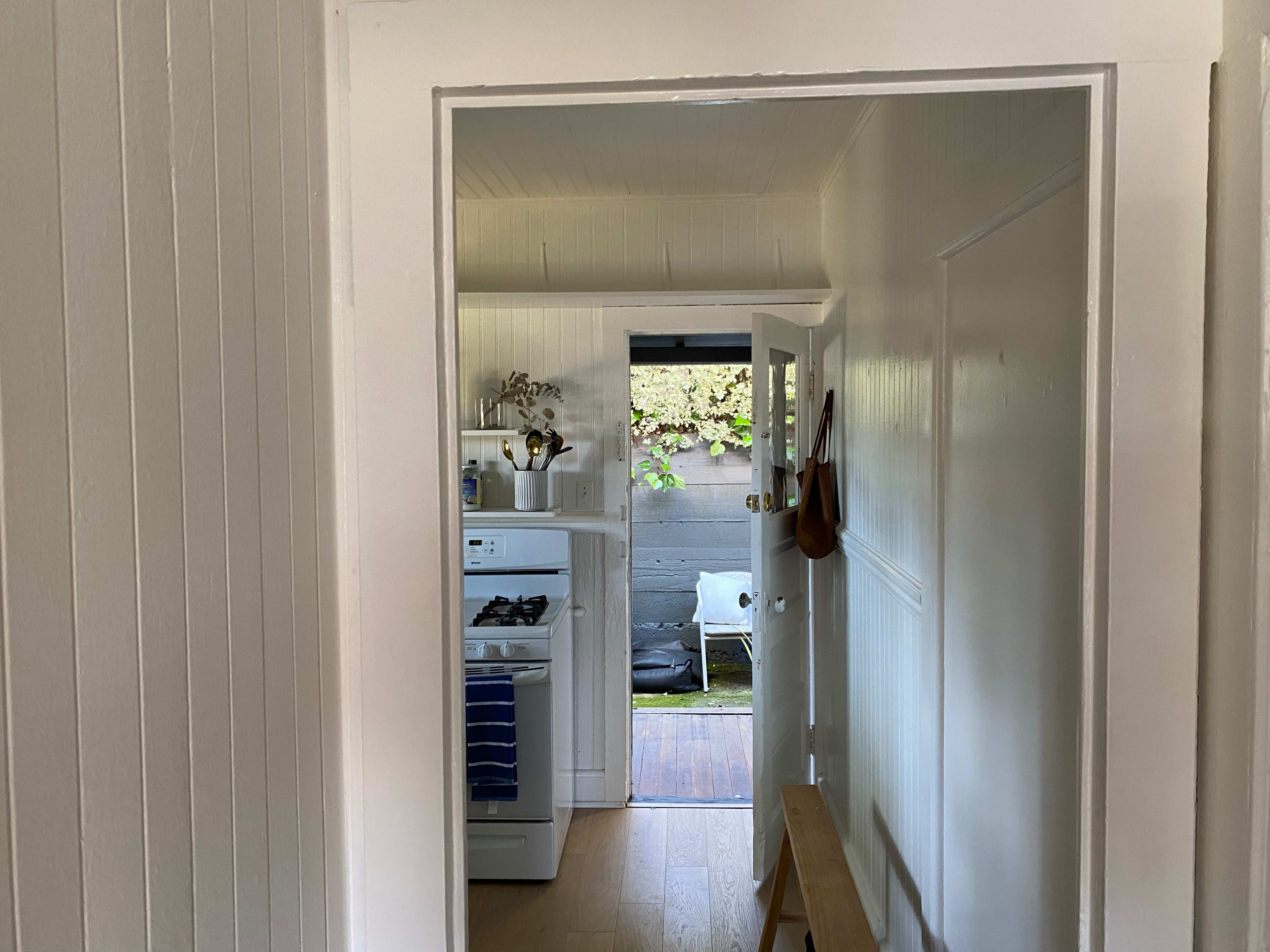 A hallway leads to a kitchen where a stove is visible, with an outdoor patio seen through an open door.