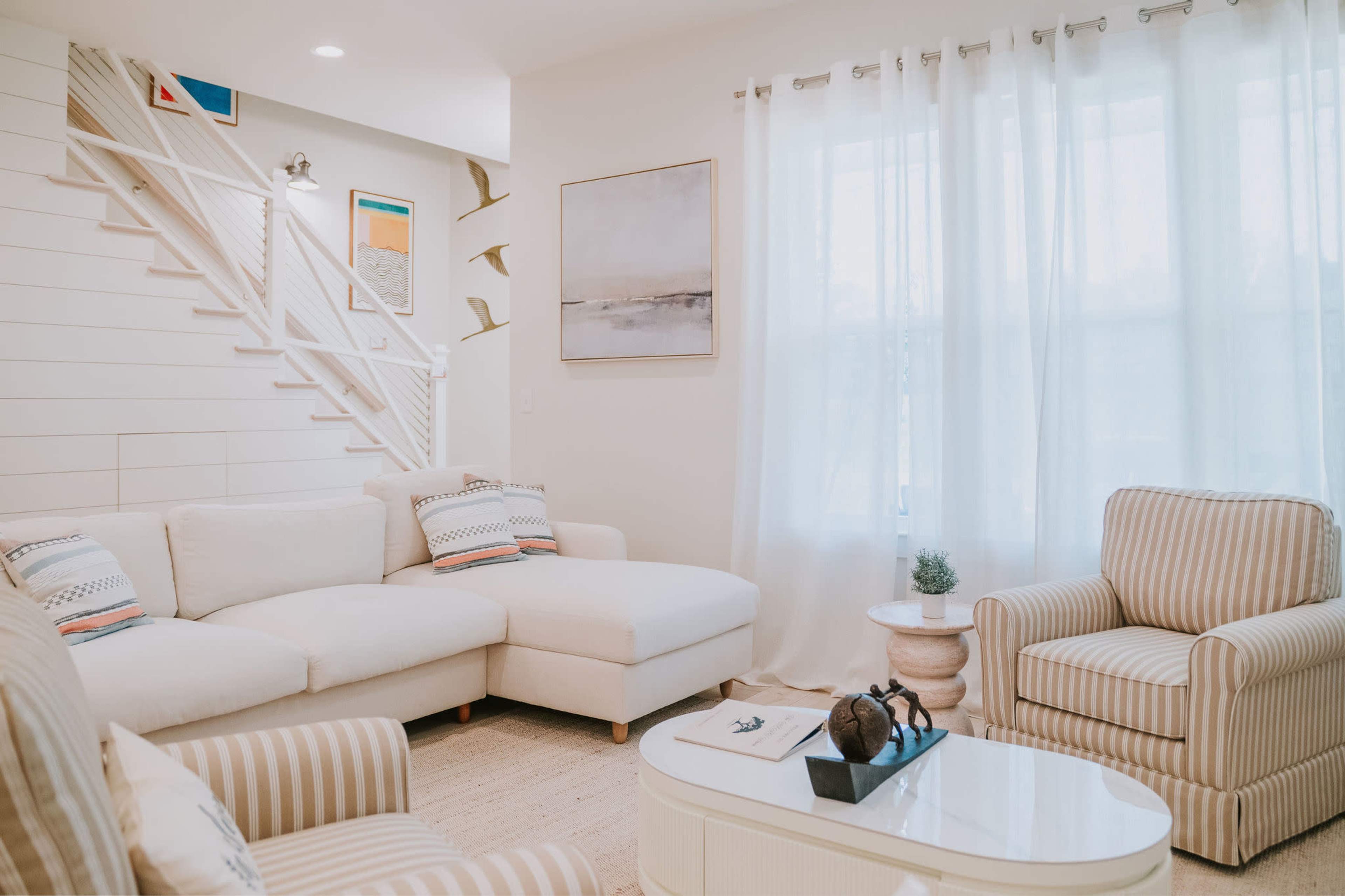 The image shows a well-lit living room with a white sectional sofa, a striped armchair, and a coffee table, featuring a staircase in the background adorned with decorative artwork.