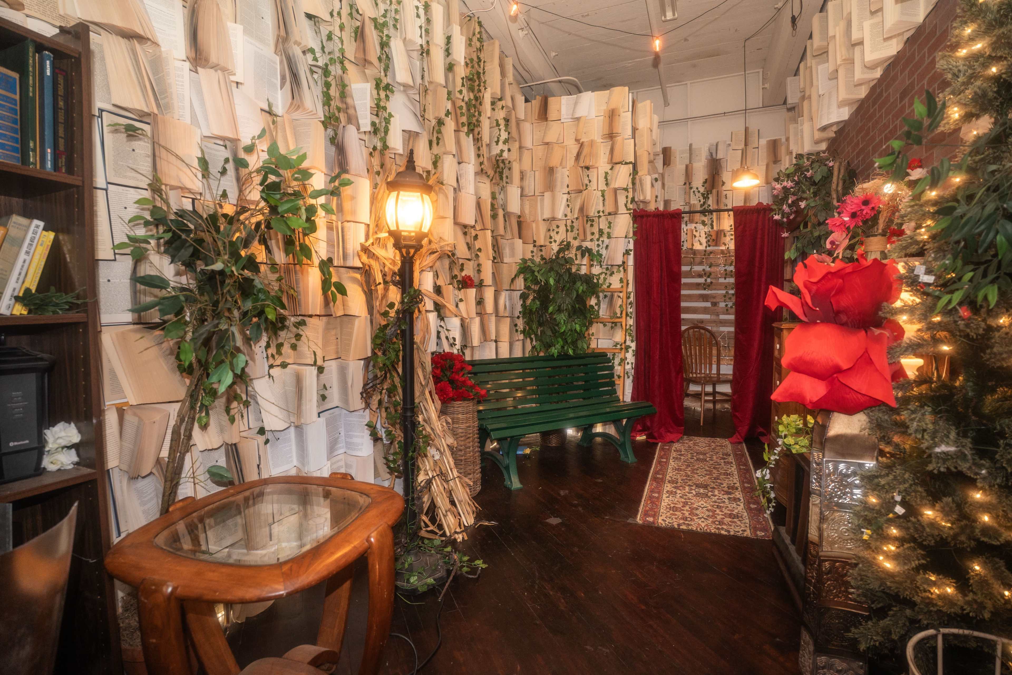 The image depicts a cozy room with walls covered in opened books, surrounded by greenery, a green bench, and decorative elements like a lamp and a Christmas tree.