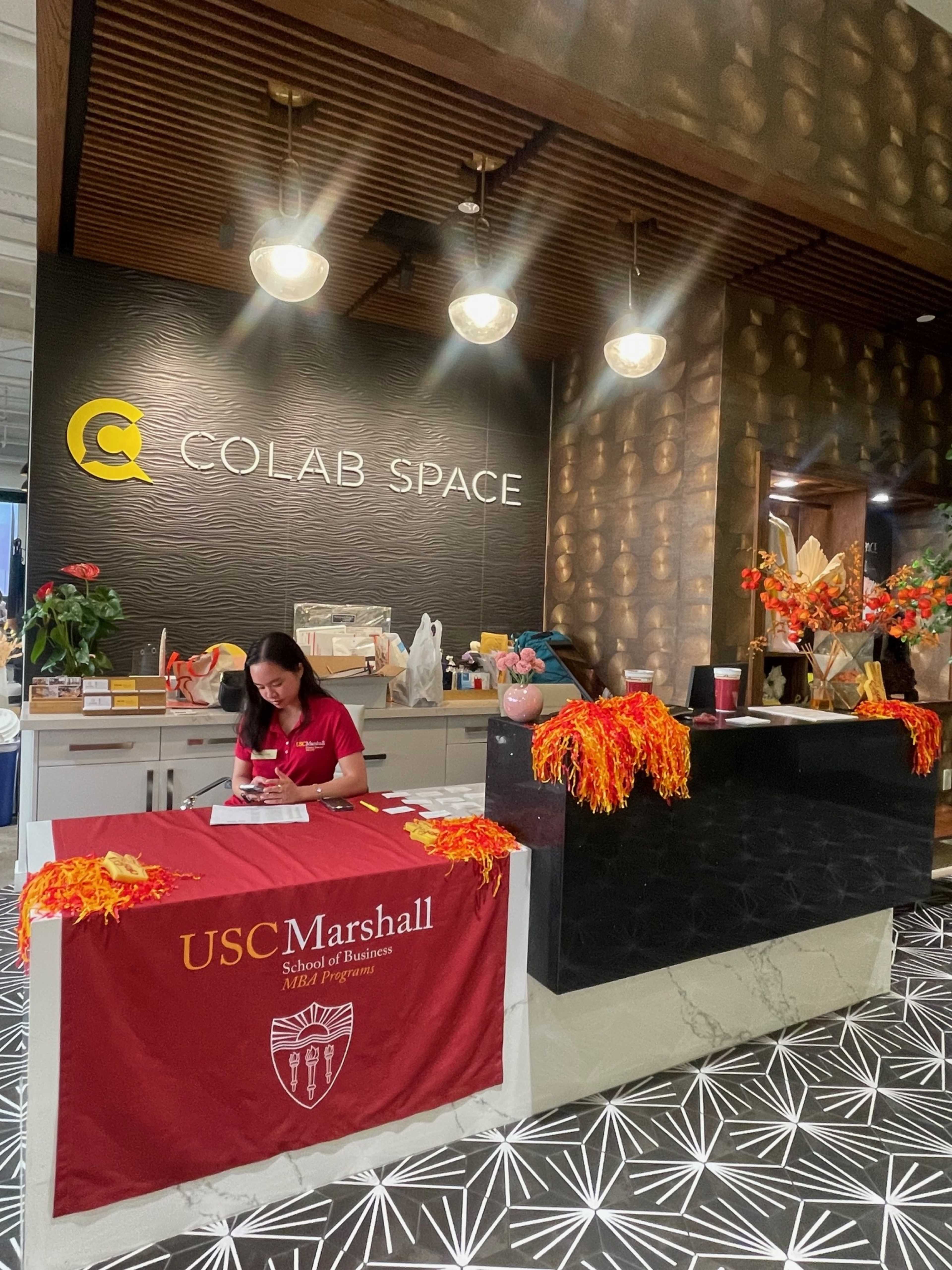 A woman sits at a reception desk in a modern workspace, surrounded by decorative plants and a USC Marshall School of Business banner.