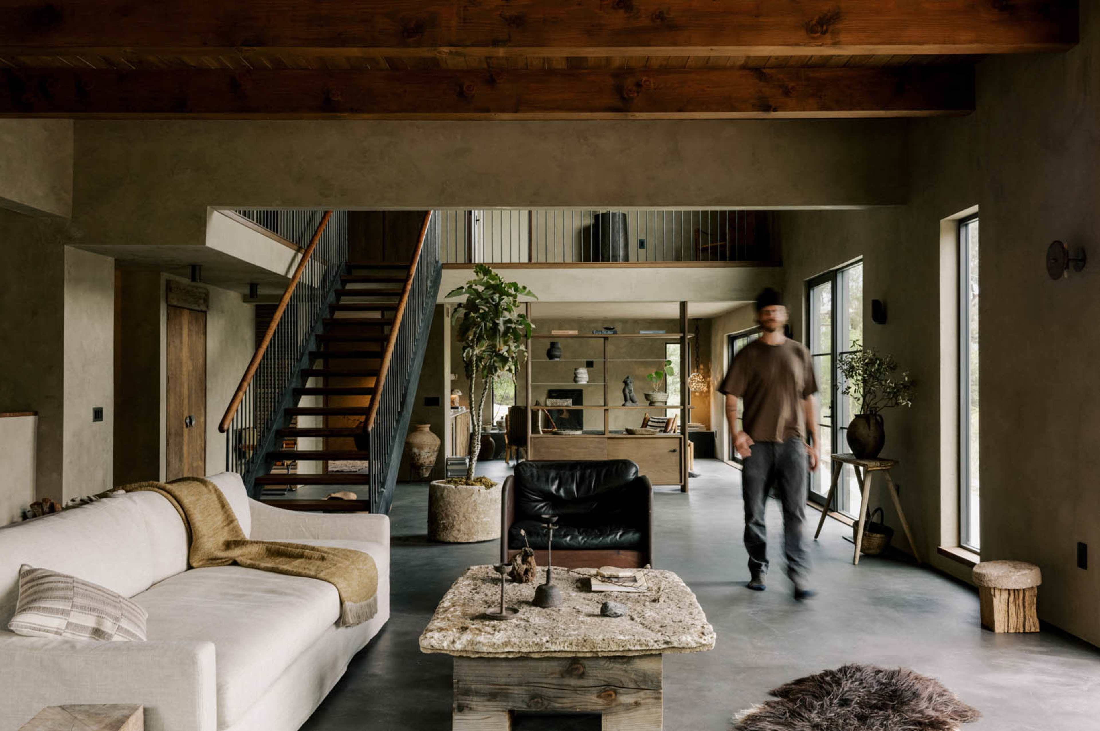 A person walks through a spacious, modern living area featuring a white sofa, dark leather chair, and a rustic coffee table.