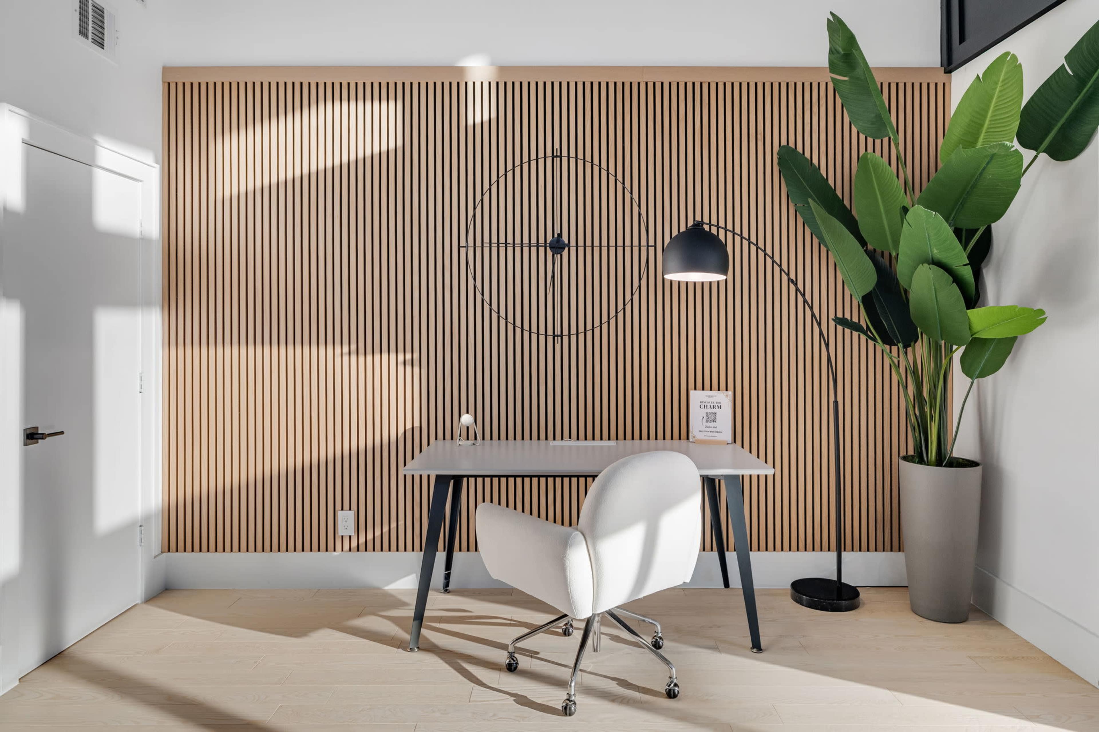 The image shows a modern office space with a wooden slatted wall, a desk with a chair, a floor lamp, and a large potted plant.