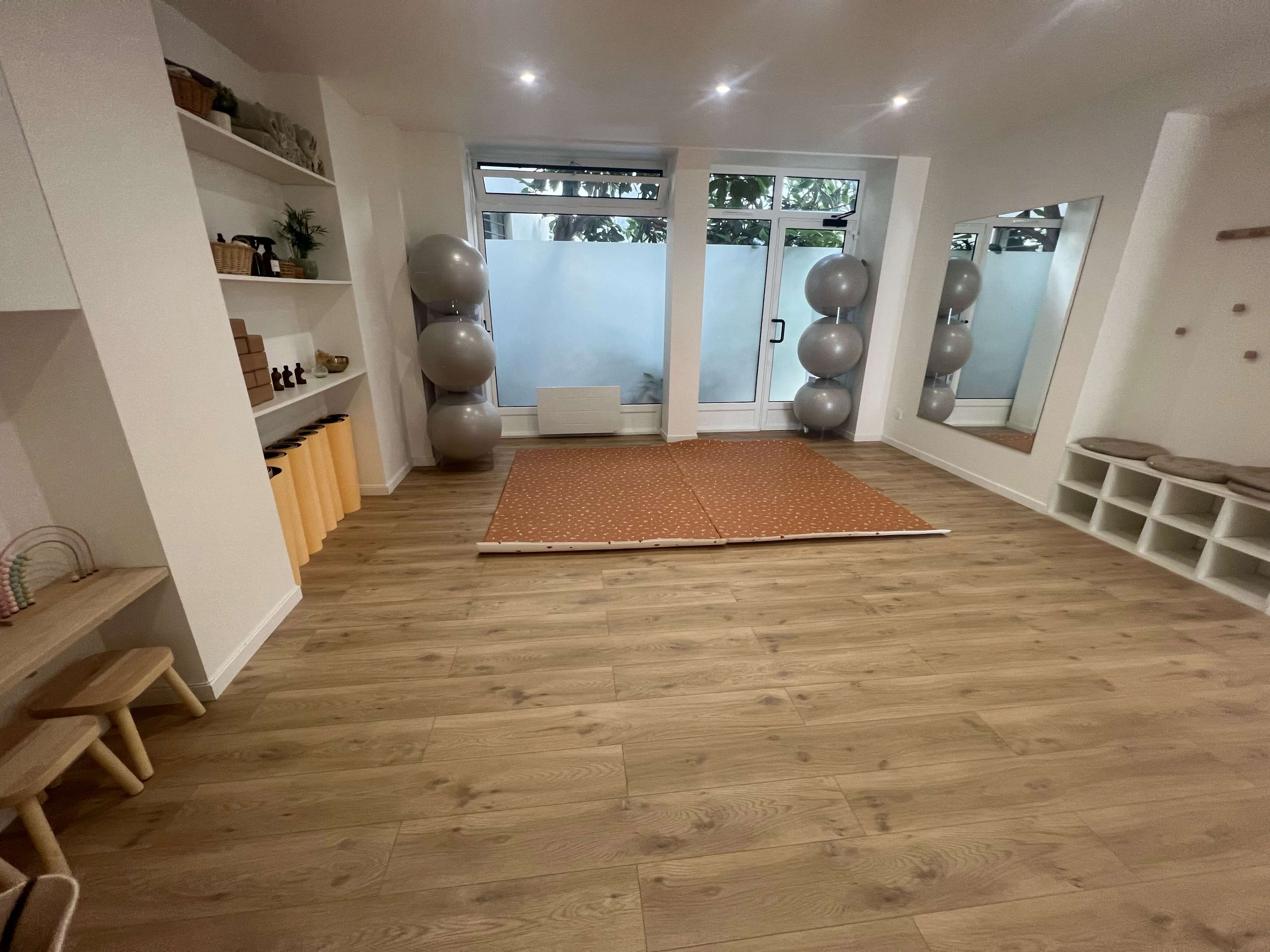 The image shows a spacious room with light wooden flooring, two large mirrors, exercise balls in a corner, and a play area with a mat and shelves.