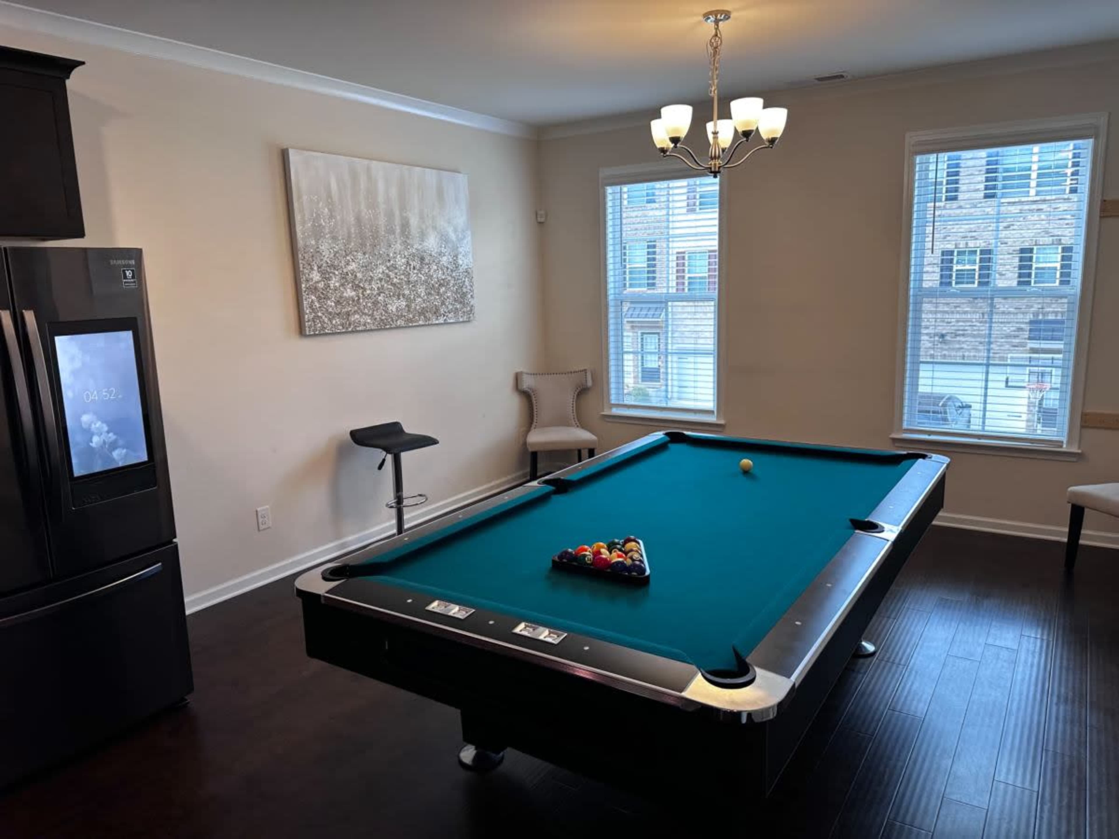 The scene shows a pool table in a light-colored room with large windows, a modern fridge, and stylish chairs.