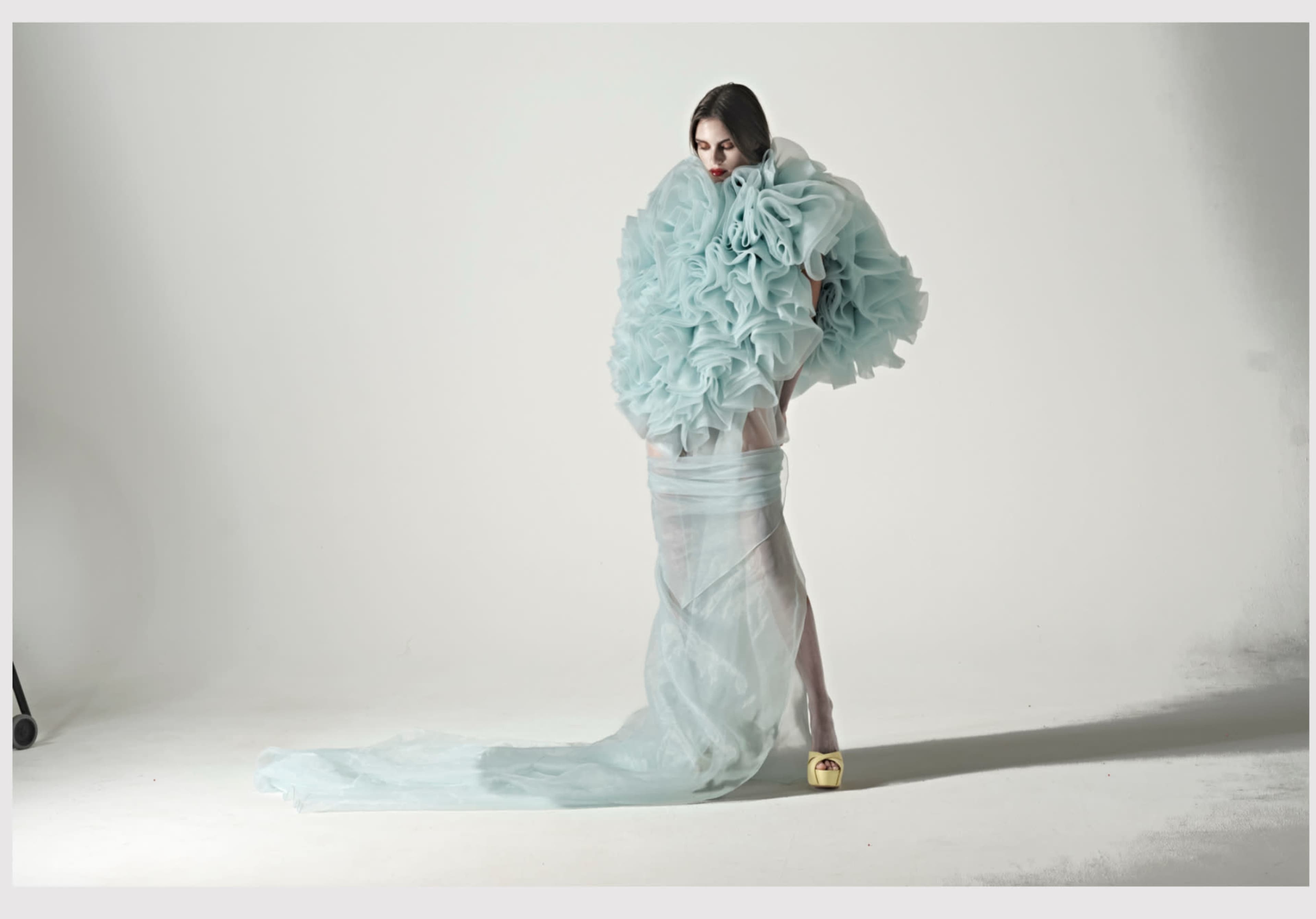A model stands in a studio, wearing an oversized light blue ruffled garment that flows to the floor.