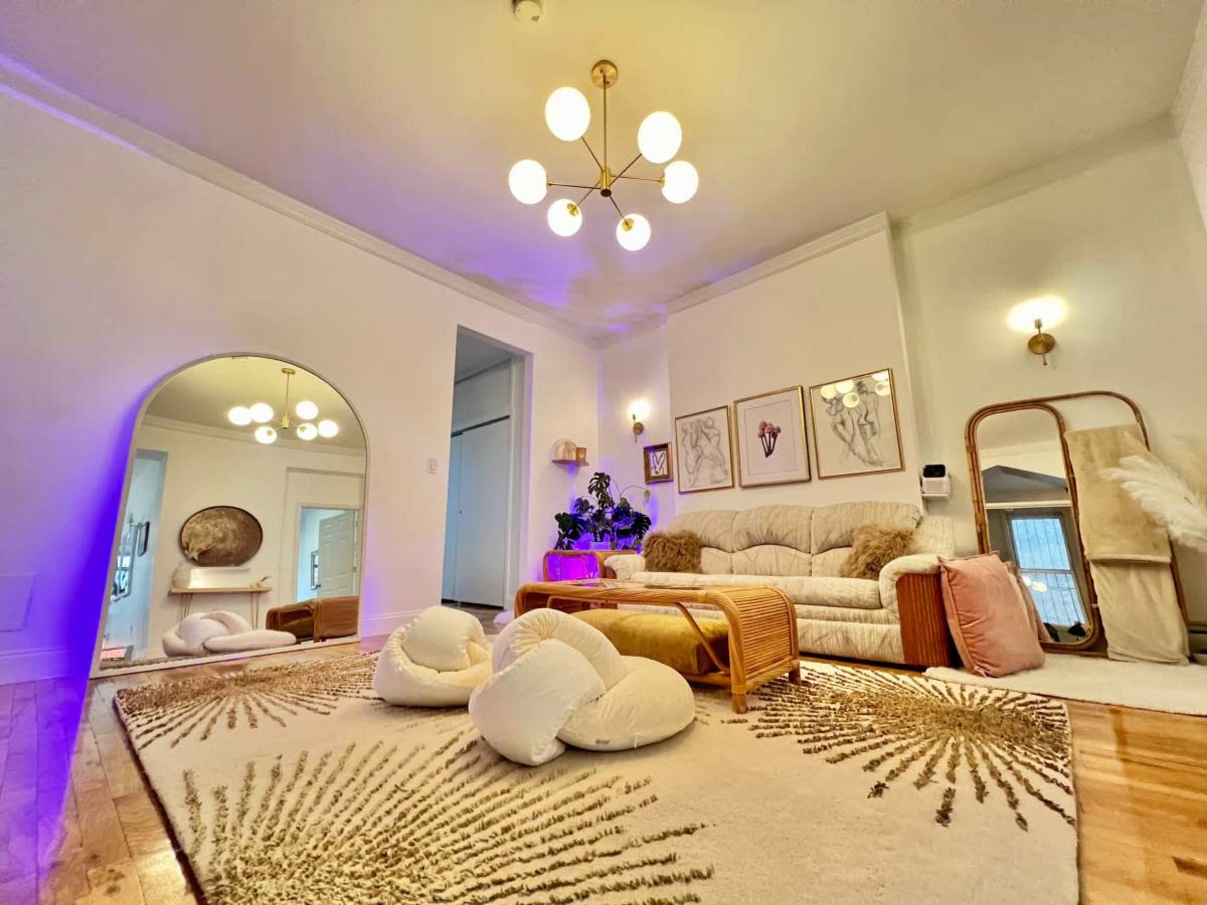 The image shows a bright living room featuring a large area rug, plush seating, framed artwork on the walls, and a decorative mirror.