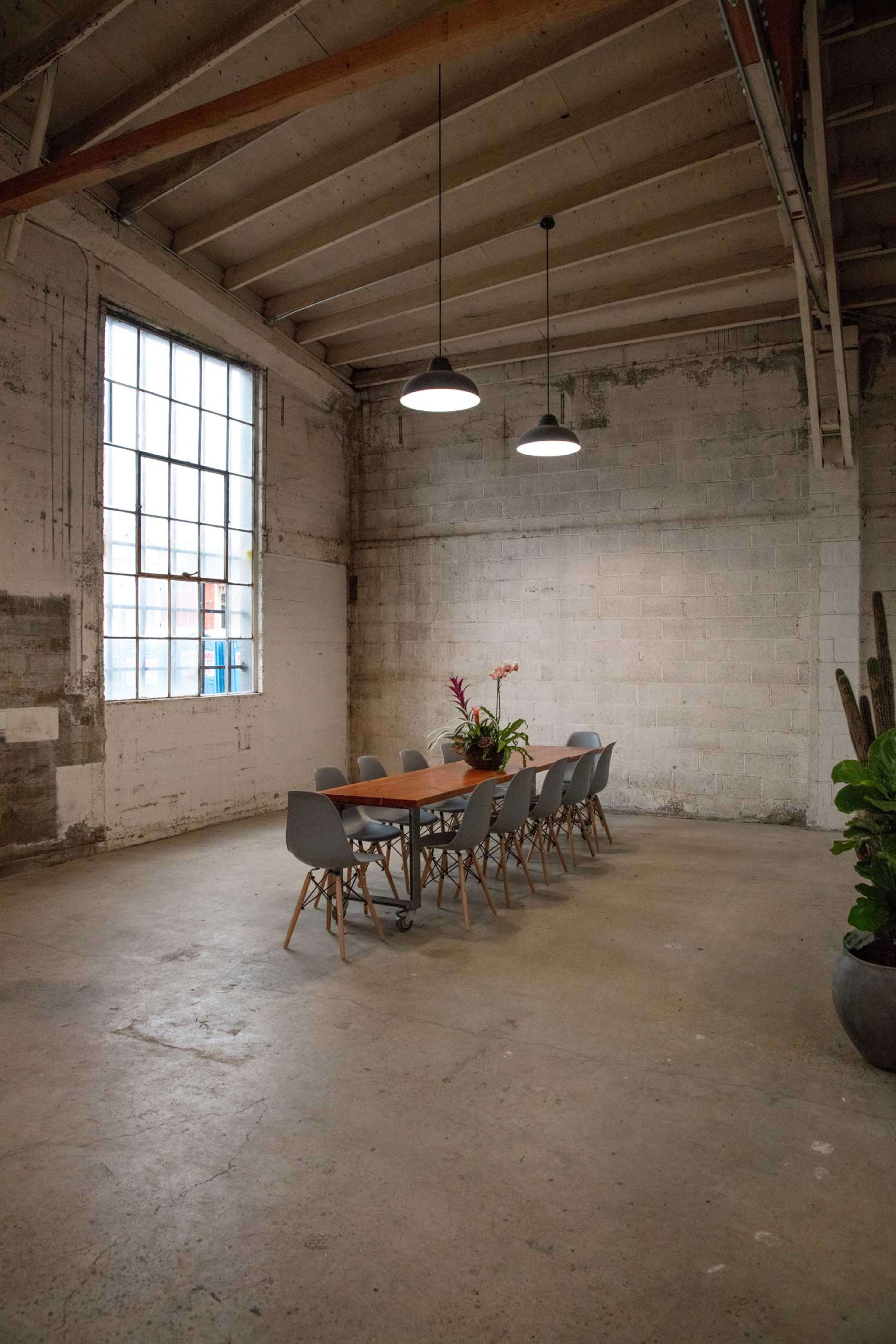 A long wooden table with chairs is set in a spacious, minimalist room with concrete walls and large windows.