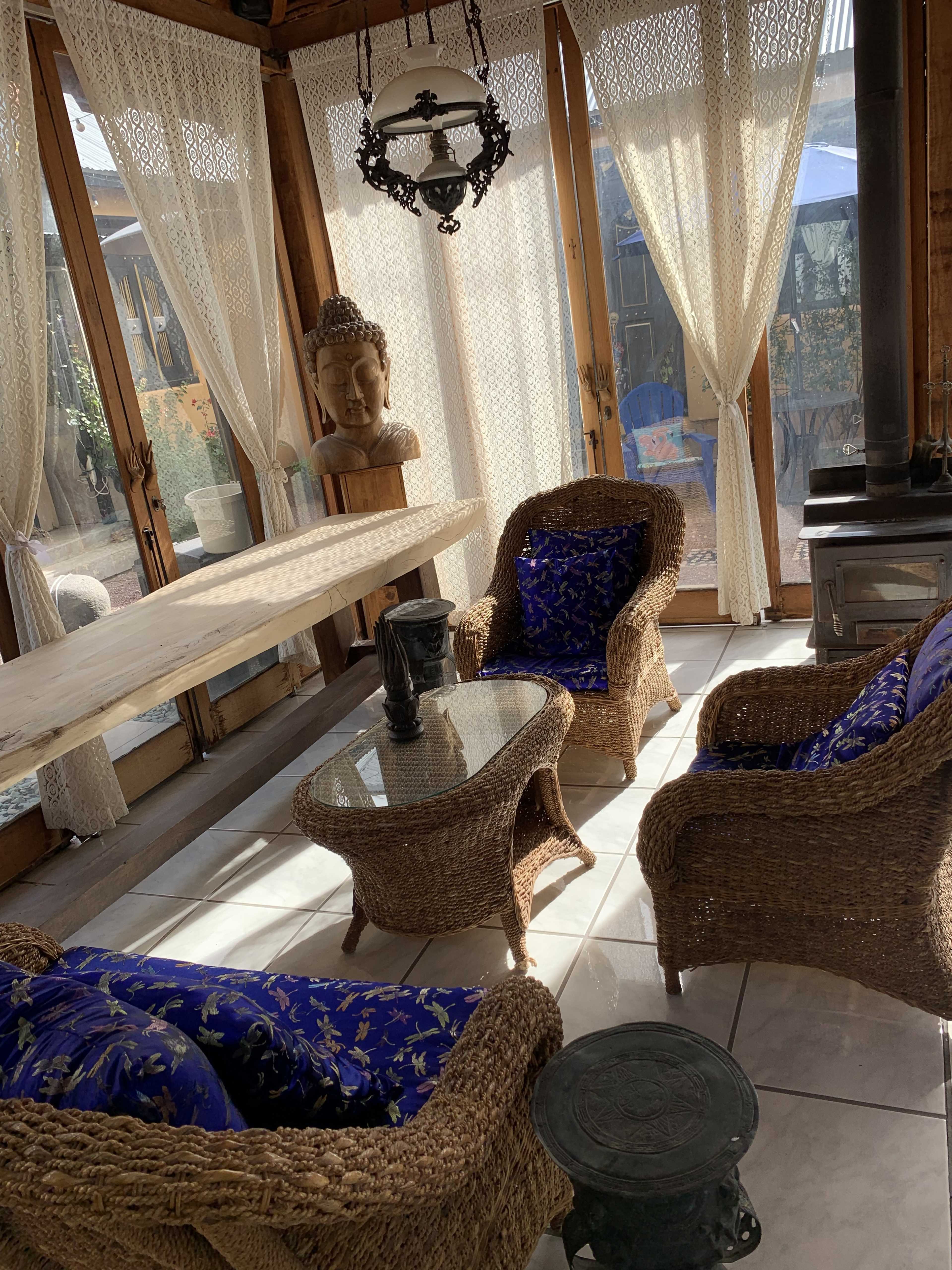 A sunlit sunroom with woven furniture, a glass table, and sheer curtains, featuring a decorative Buddha head and a stove.