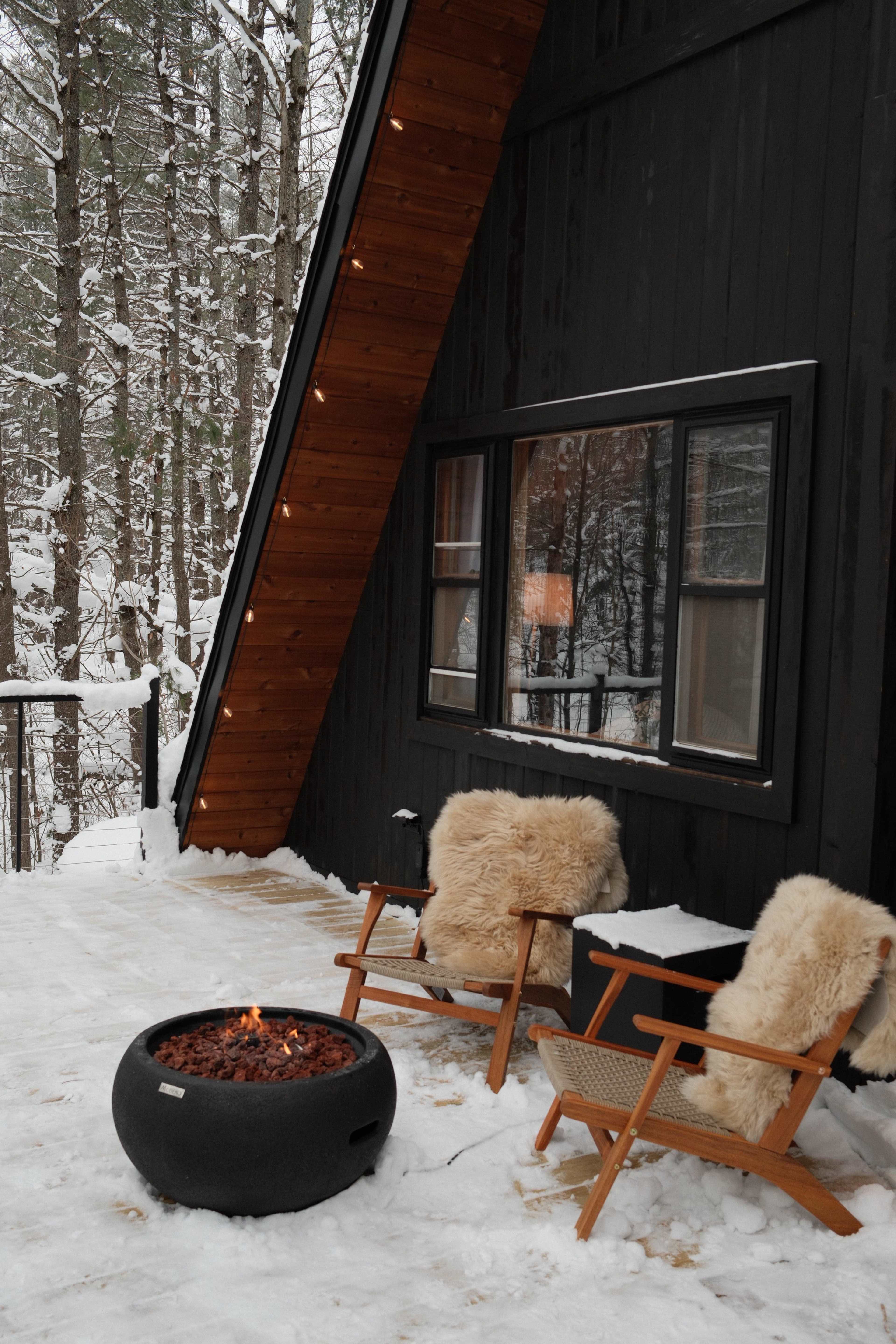 A black A-frame cabin, surrounded by snow-covered trees, features two deck chairs with fur cushions positioned around a circular fire pit.