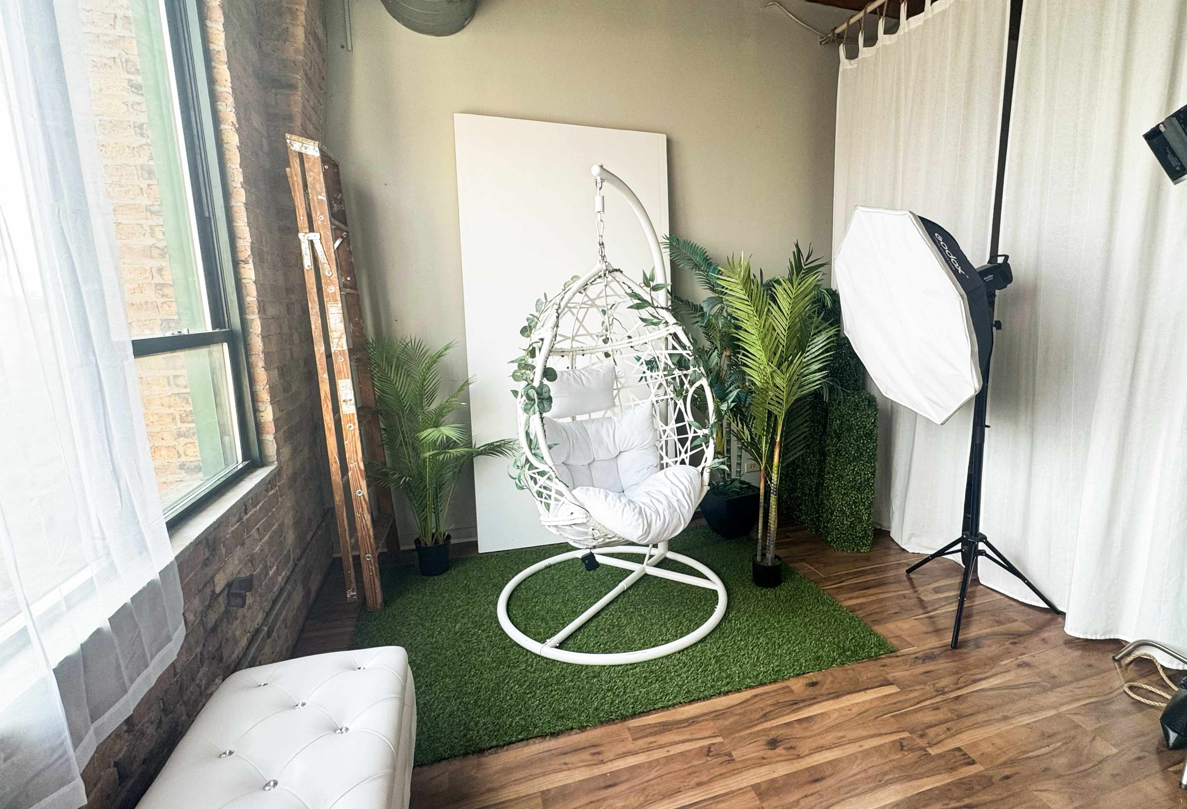 The image shows a cozy studio space featuring a white hanging chair surrounded by potted plants, a faux grass rug, and photography lighting equipment.