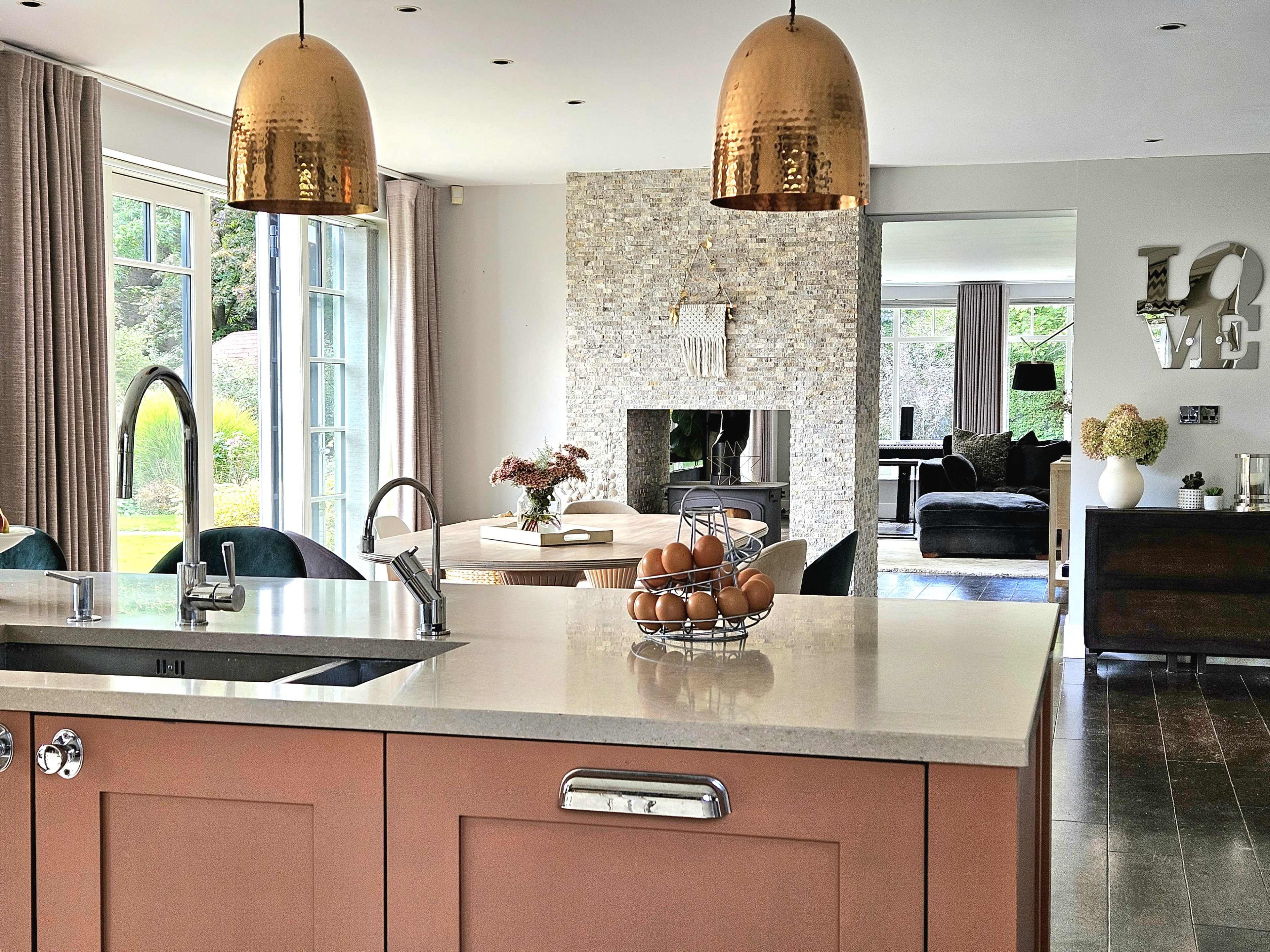 A modern kitchen features a central island with a sink and a bowl of eggs, complemented by copper pendant lights, a stone fireplace, and a view into a cozy living area.