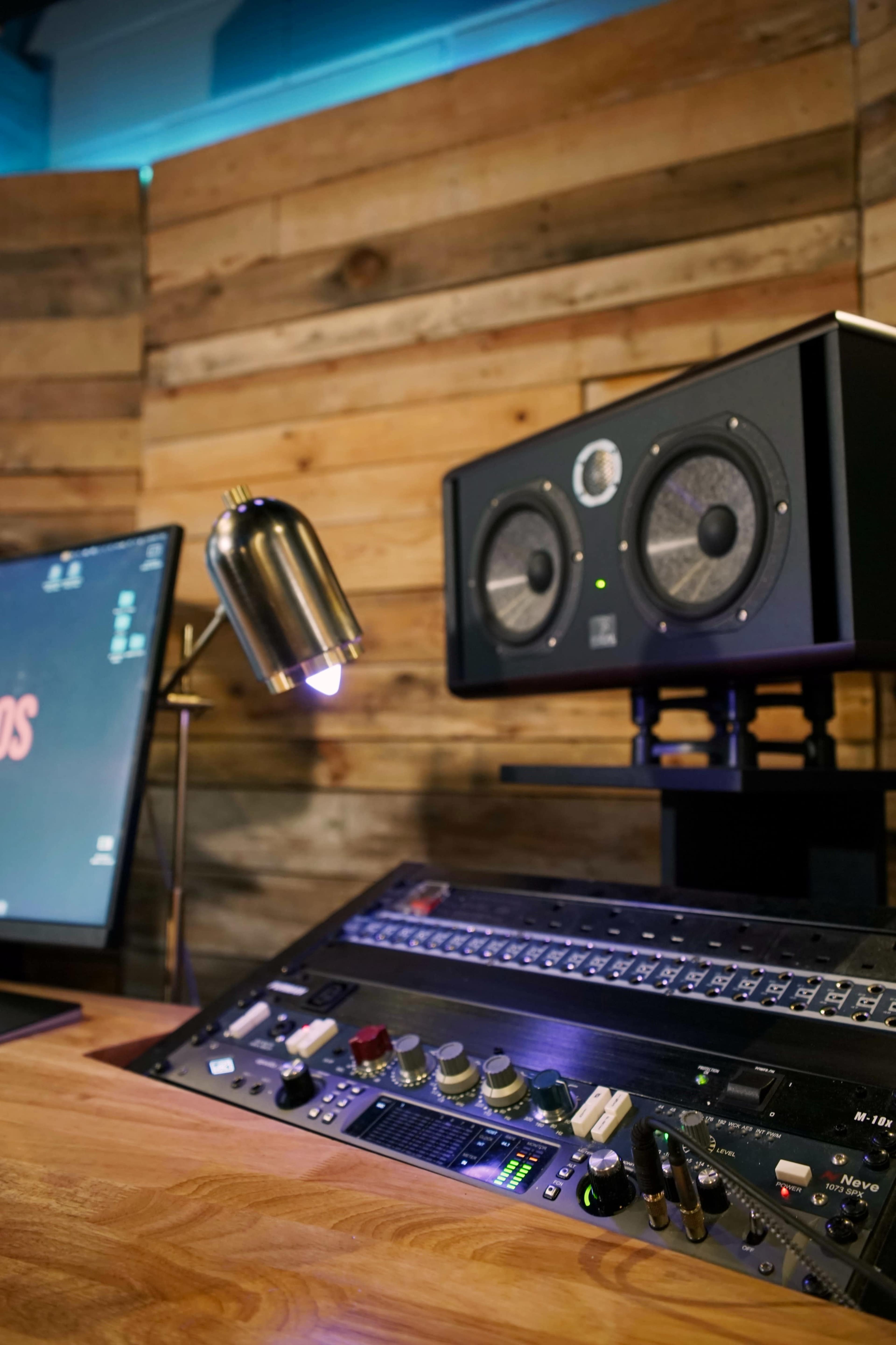The image shows a modern audio control setup with a mixing console, studio monitors, a computer screen, and a desk lamp against a wooden backdrop.