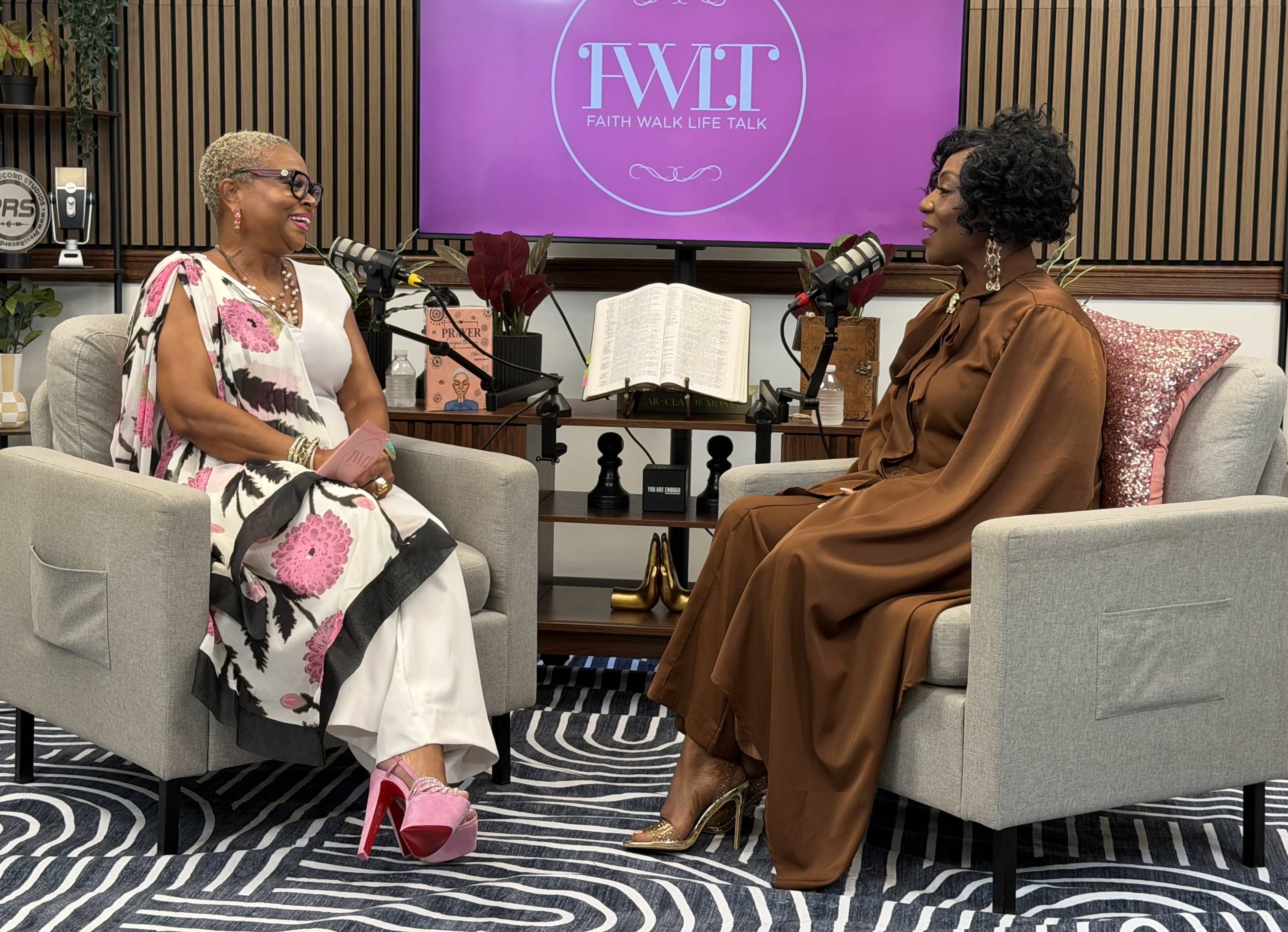 Two women are seated on modern armchairs in a studio setting, engaged in conversation with a backdrop displaying the title "Faith Walk Life Talk."