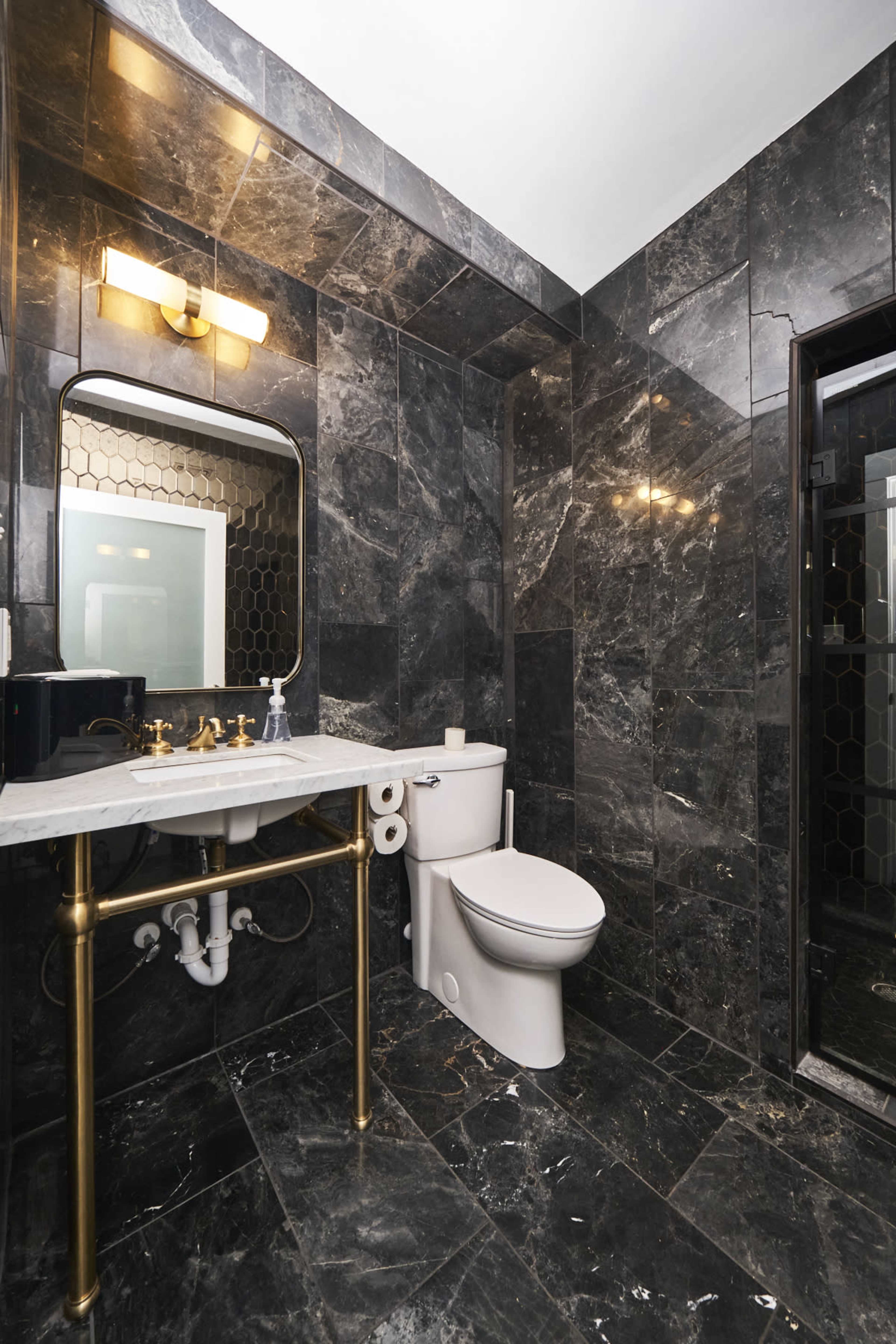 The bathroom features dark marble walls, a white toilet, a gold-accented sink with a mirror, and a glass-enclosed shower.