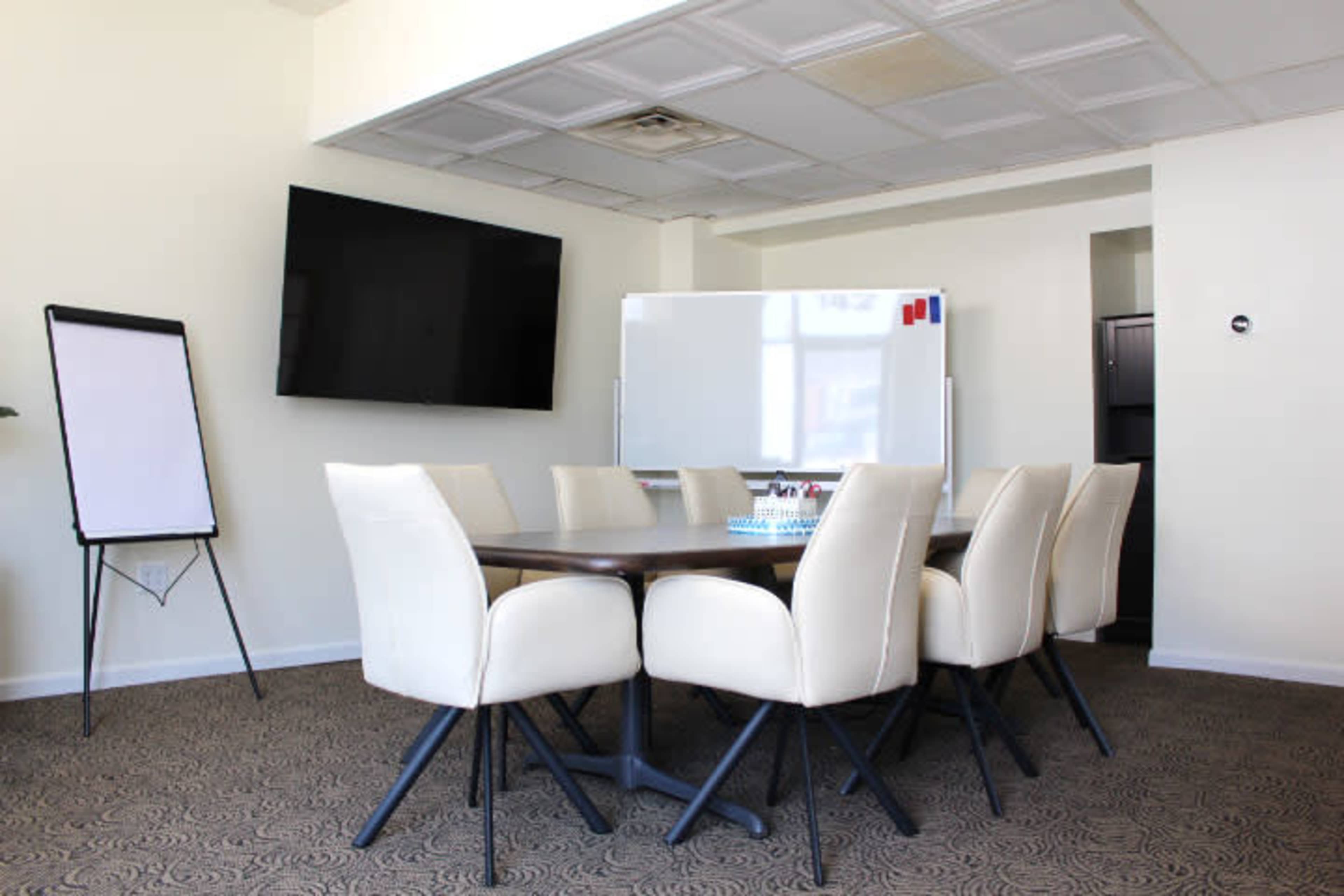 A conference room features a large table surrounded by eight cream-colored chairs, with a whiteboard and a television mounted on the wall.