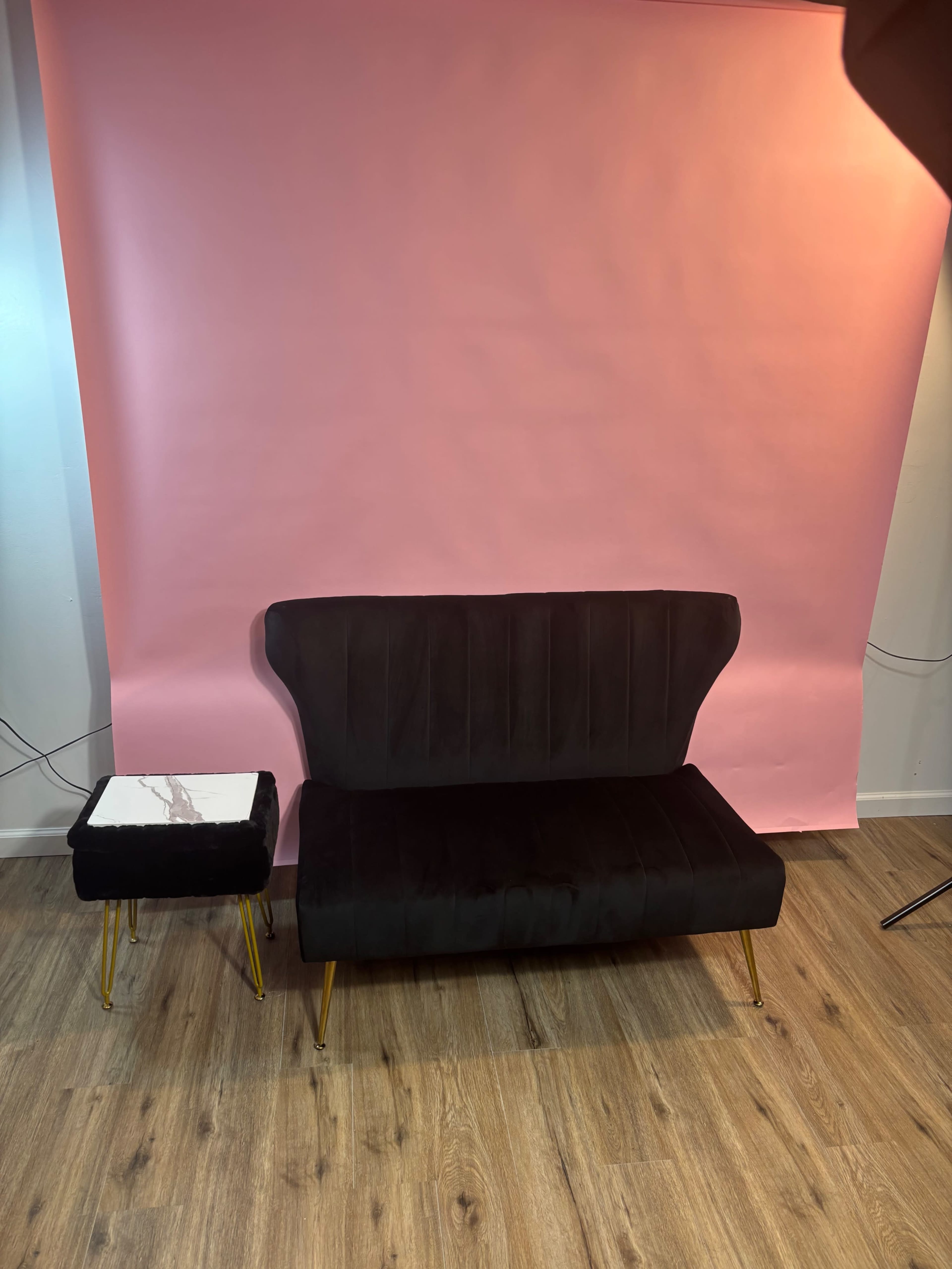 A black velvet loveseat with gold legs is positioned next to a small, square side table featuring a marble top, against a pink backdrop.