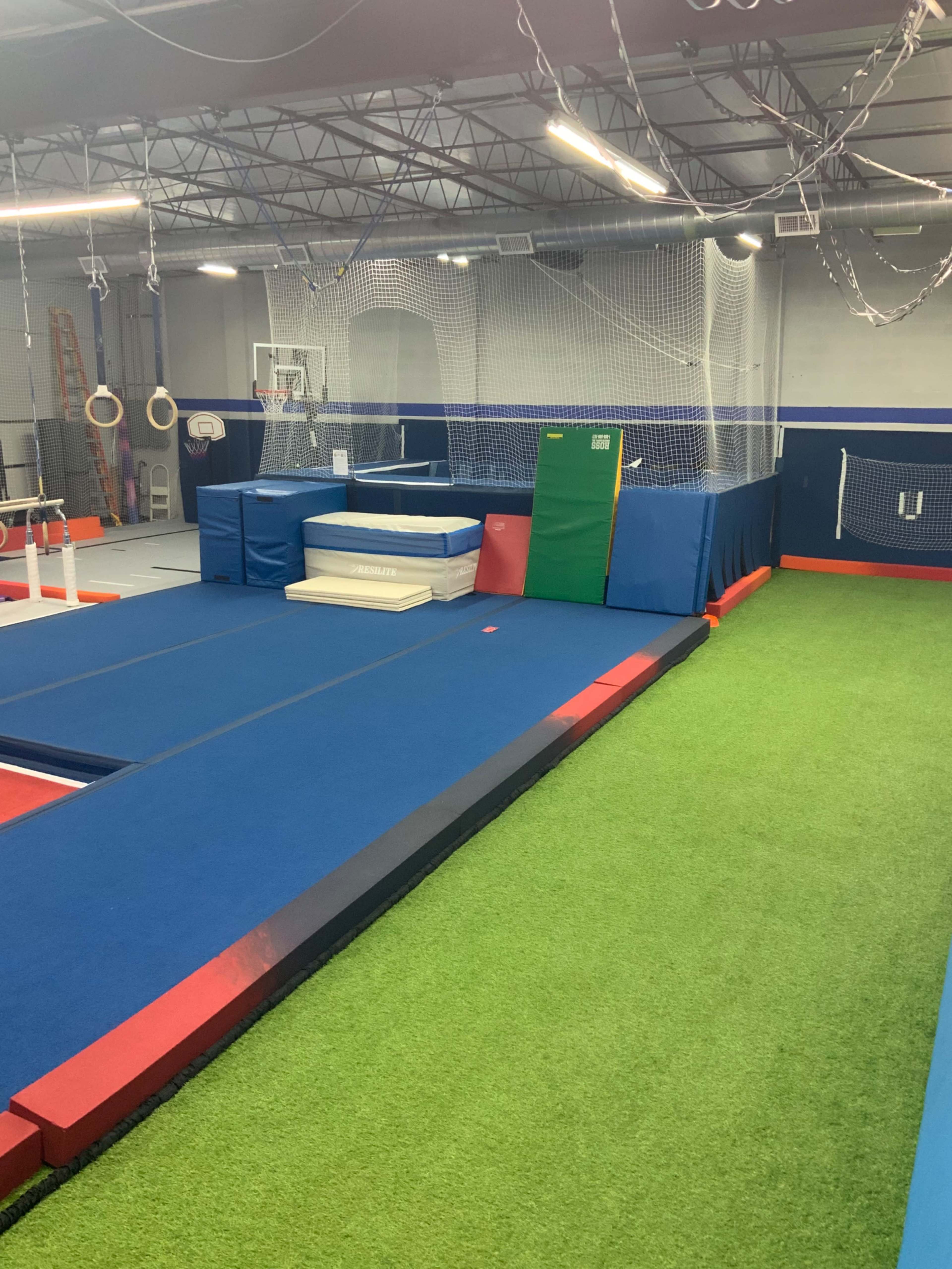 The image shows a gymnastics training area with padded surfaces, mats, and a green turf section, along with various gymnastic equipment and safety nets.