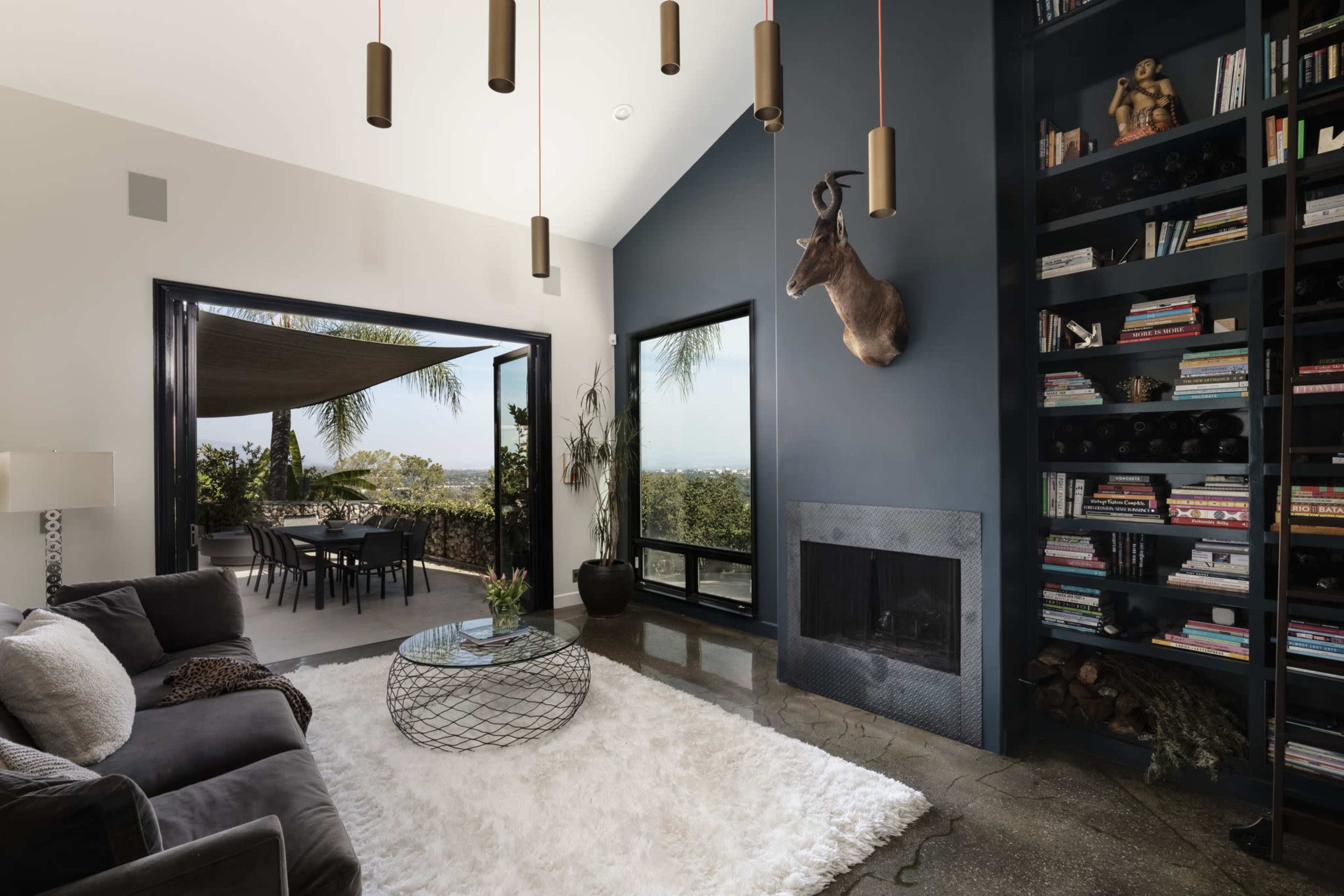 The living room features a modern design with a large bookshelf, a plush rug, a cozy seating area, and floor-to-ceiling windows that open to an outdoor dining space.