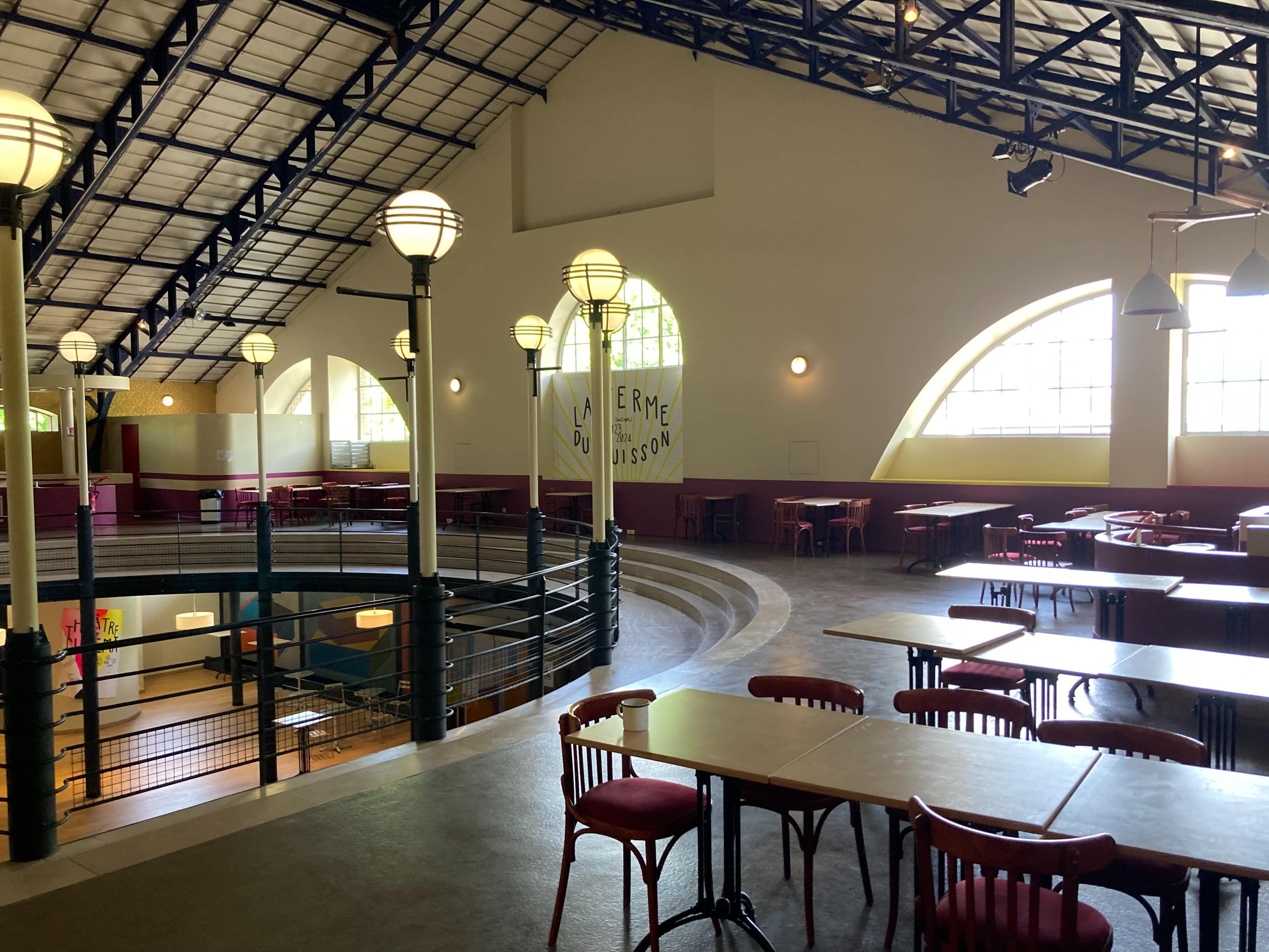 The image shows a spacious cafeteria with high ceilings, arched windows, and rows of tables and chairs arranged throughout the area.