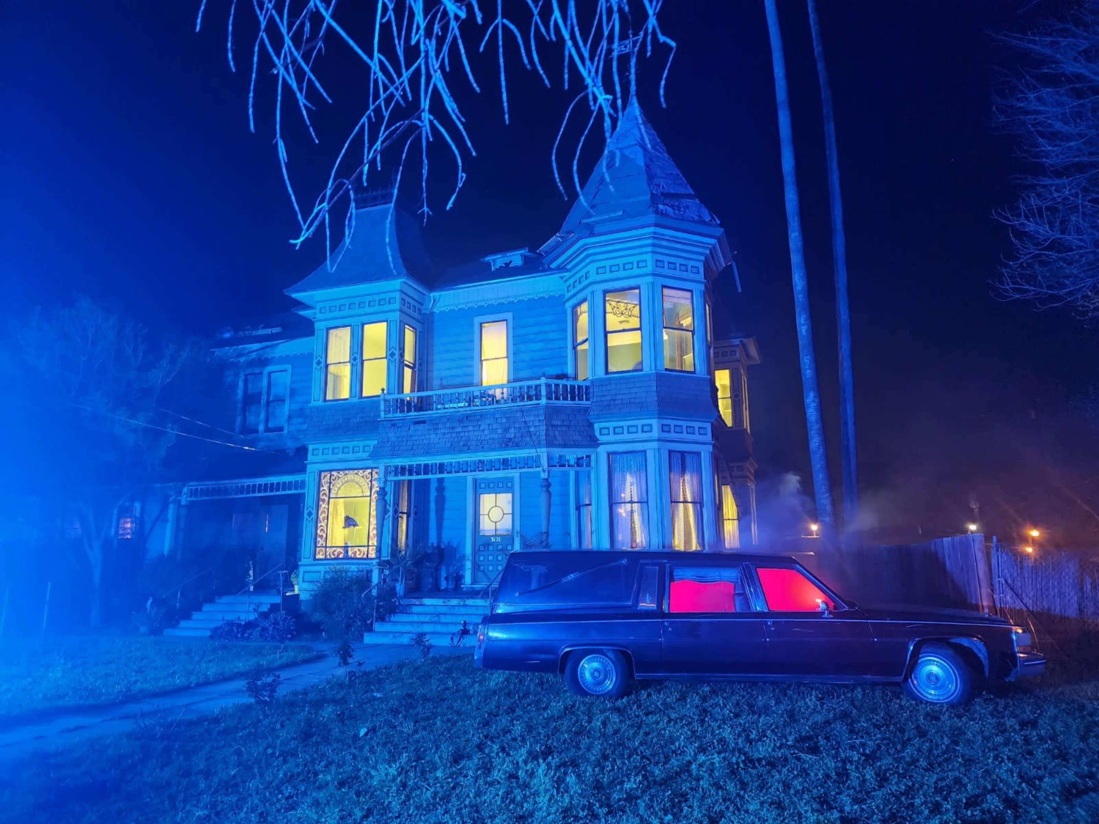 A Victorian-style house is illuminated by blue lights, with a classic hearse parked in front and the windows glowing in yellow.