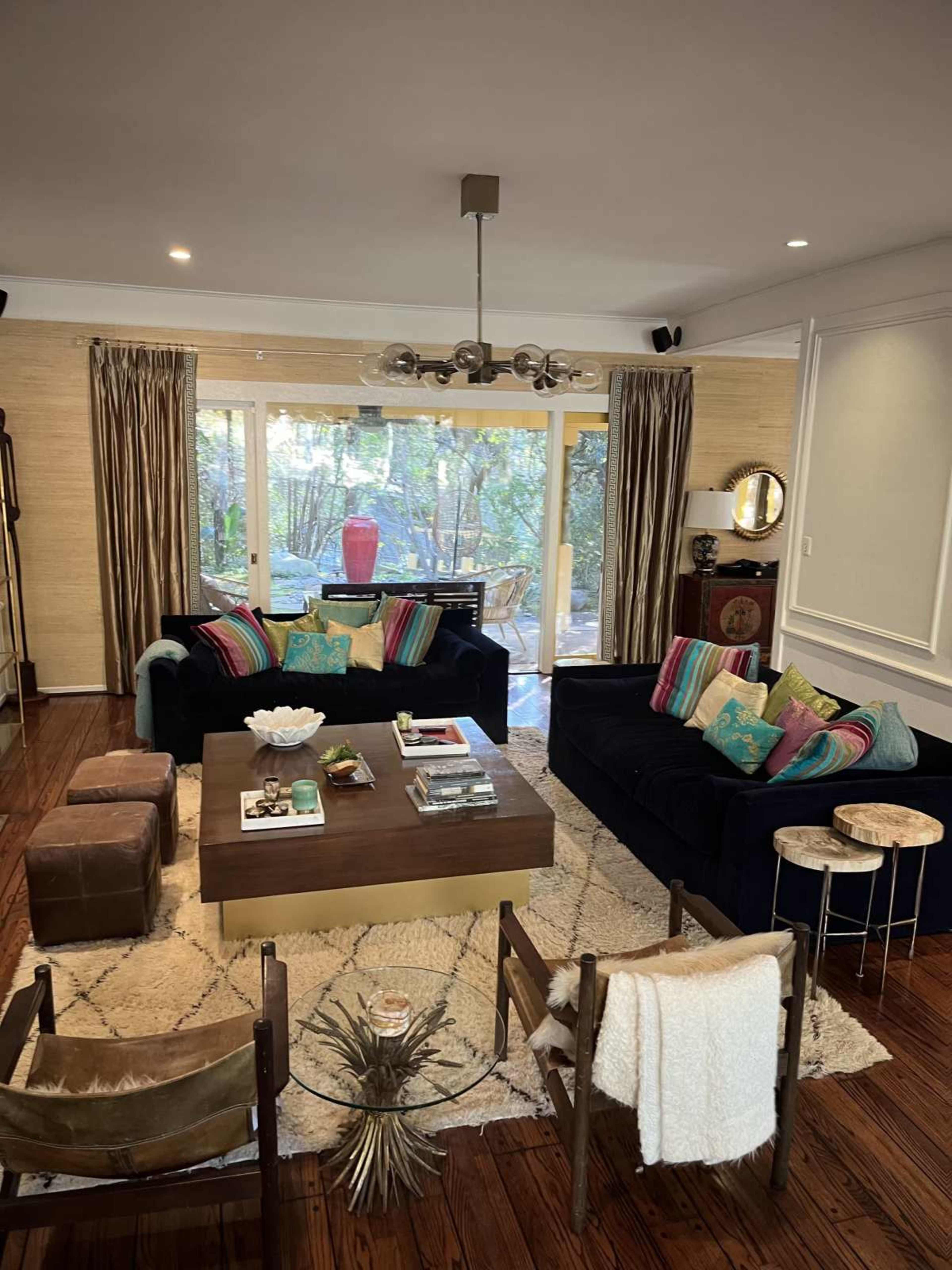 The living room features a dark lounge area with vibrant cushions, a wooden coffee table in the center, and large windows that offer a view of greenery outside.