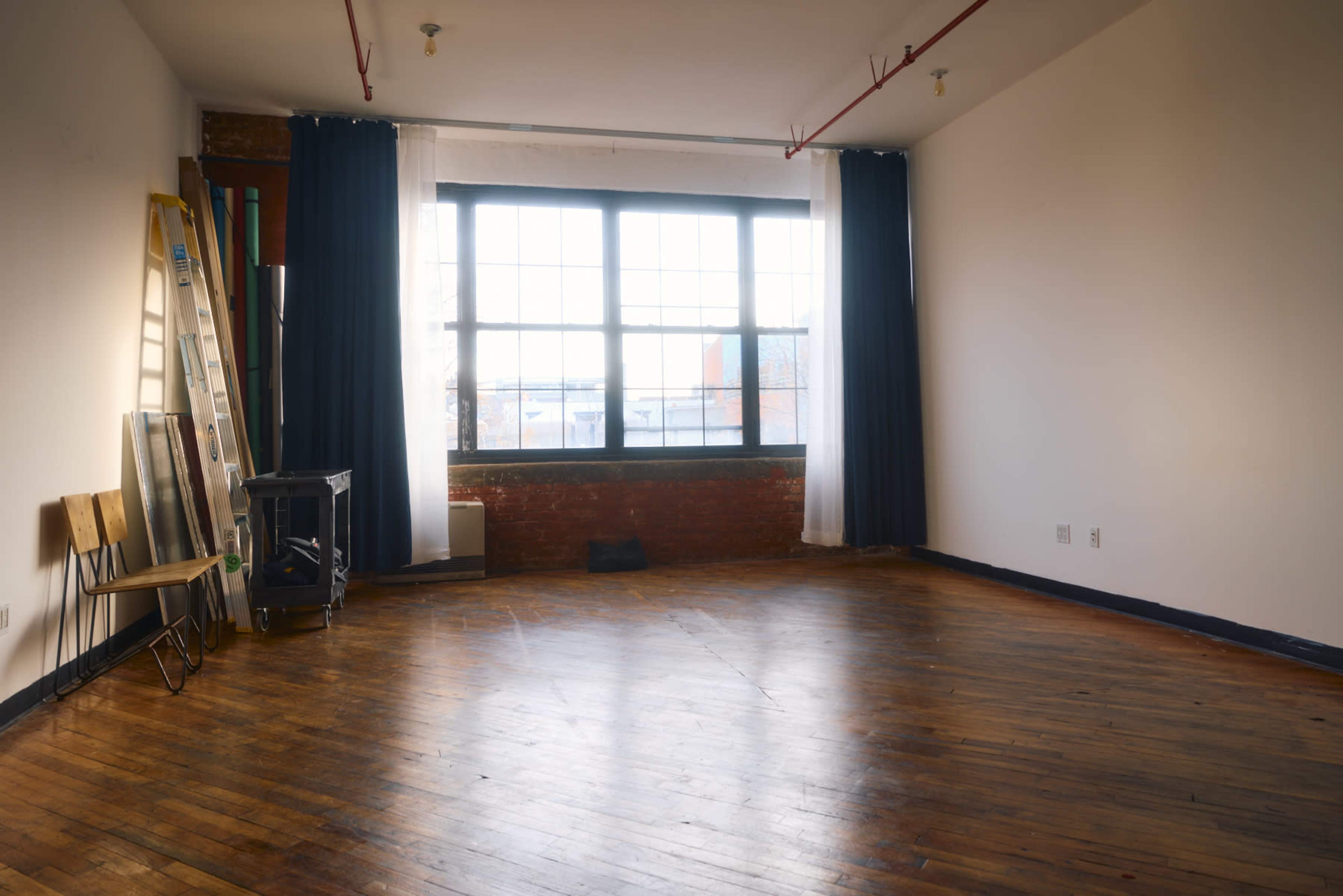 An empty room with wooden floors and large windows, featuring a few chairs, a ladder, and a small stove.