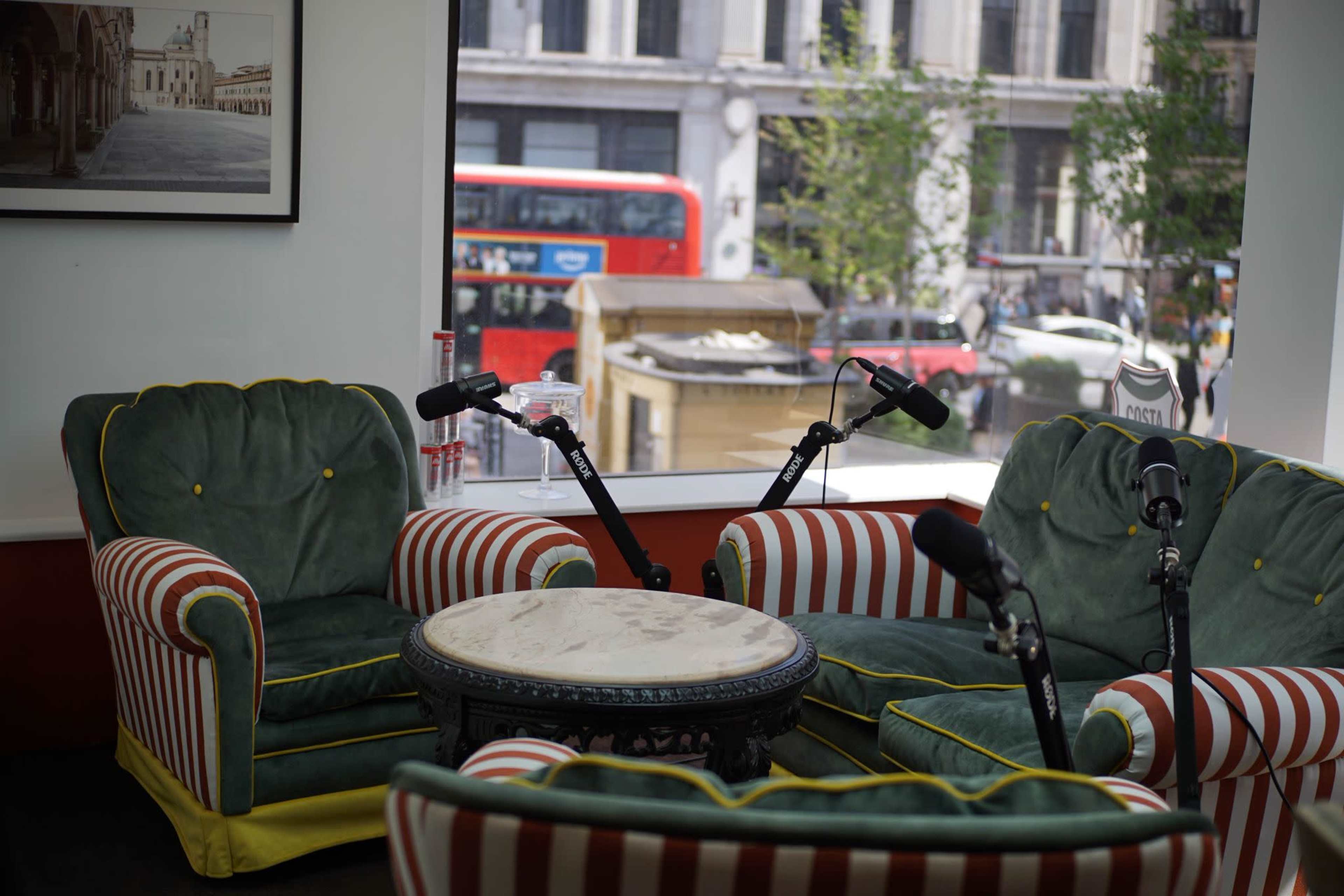 The image shows a seating area with green and striped armchairs and a round table, adjacent to a large window overlooking a busy street with a red double-decker bus.